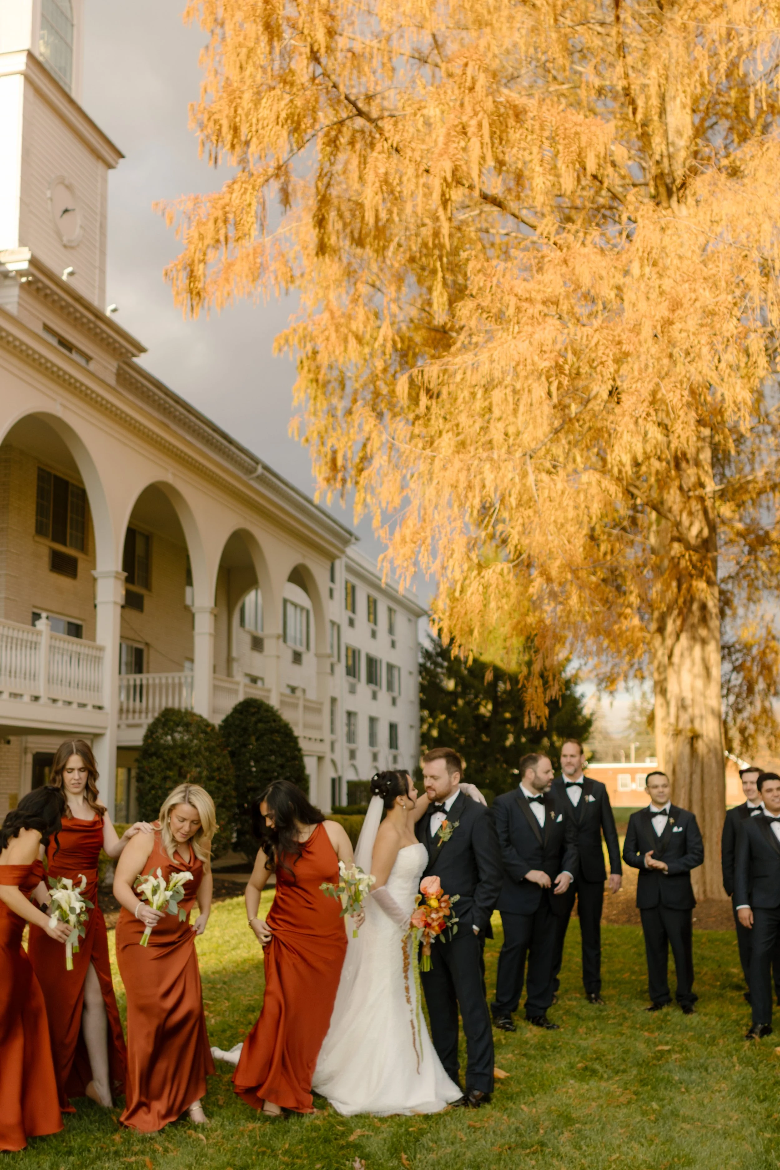 Wedding party gathers for portraits beneath golden autumn trees outside a historic estate venue.