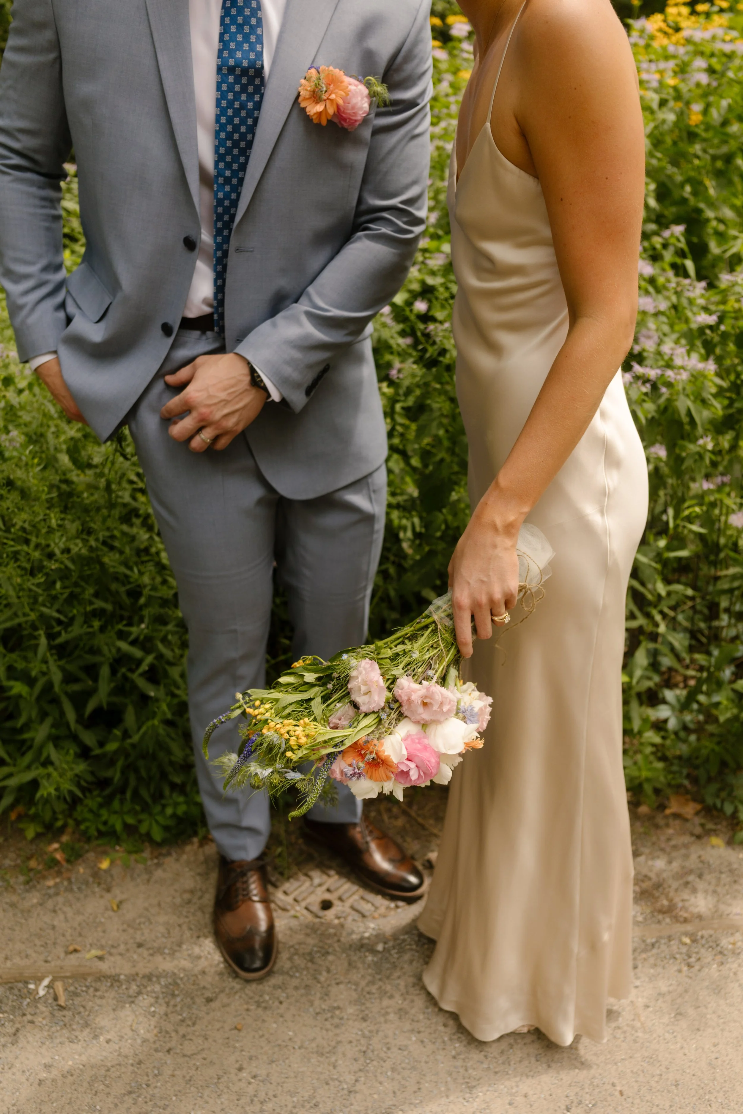 Close-up of the bride's silk wedding dress, colorful bouquet, and groom's light blue suit during an intimate garden wedding.