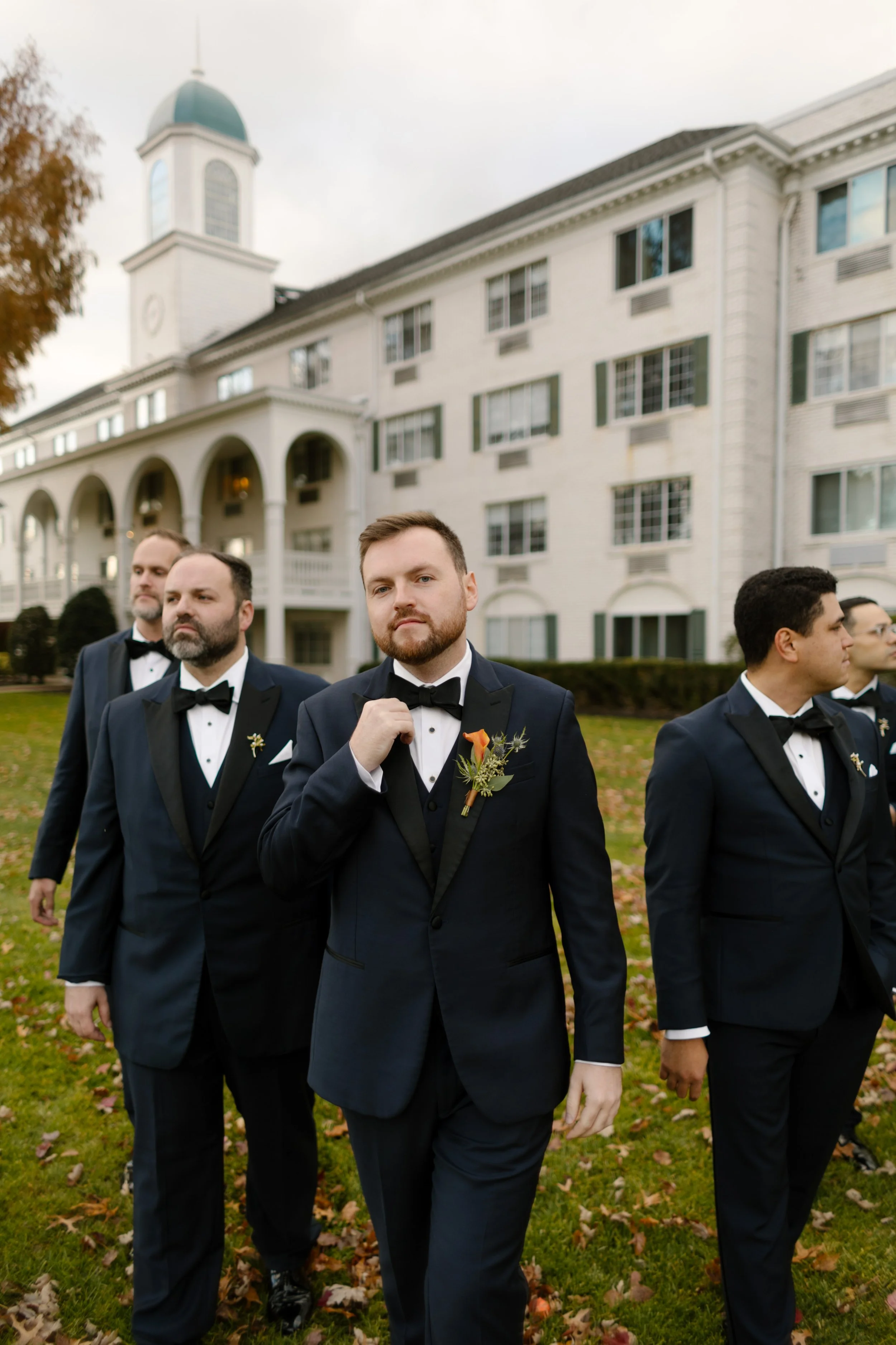 Groom adjusts his bow tie while walking with his groomsmen across the lawn at their elegant estate wedding venue.