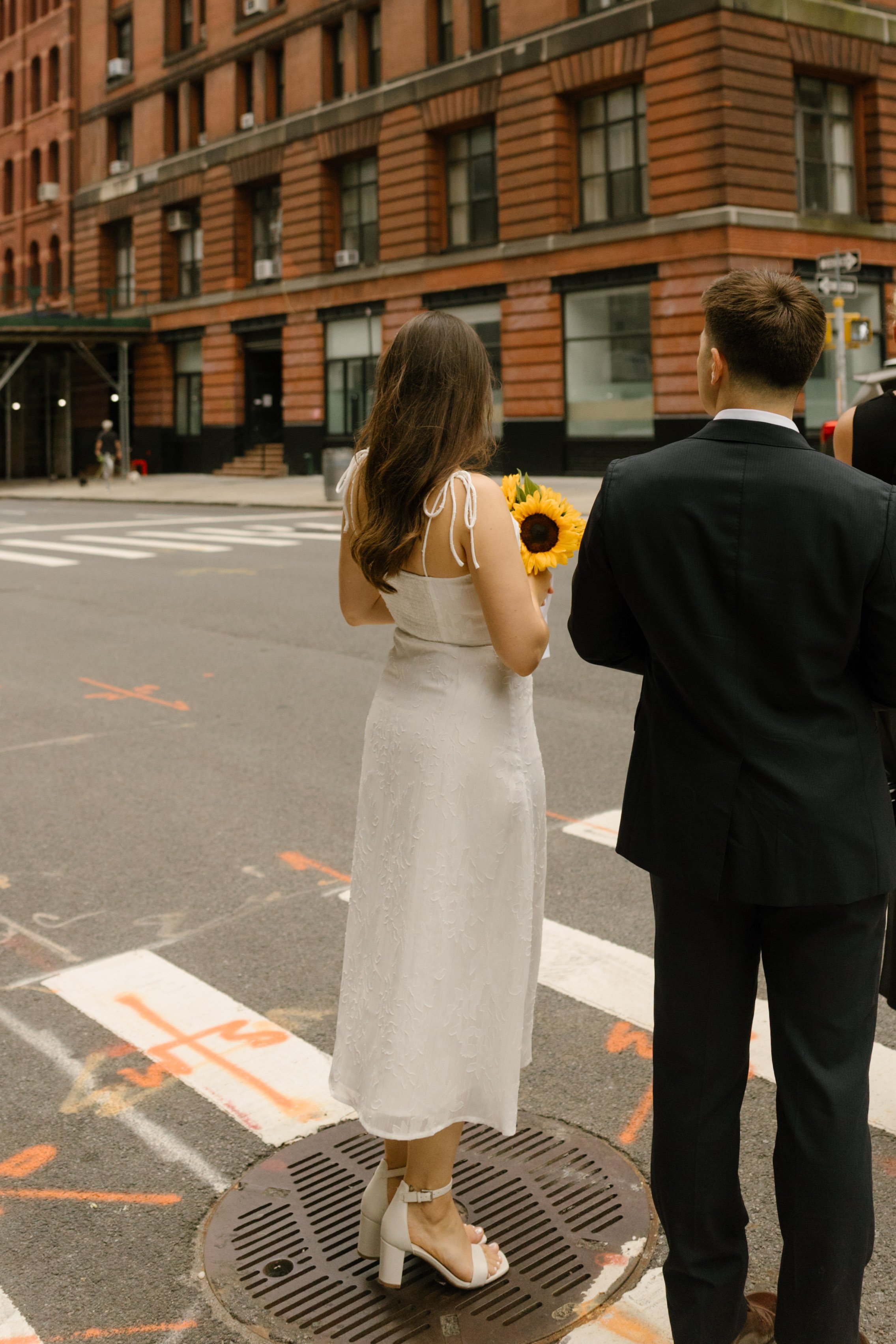 Newlyweds wait together at a Manhattan crosswalk, the bride holding a sunflower bouquet after their city wedding ceremony.