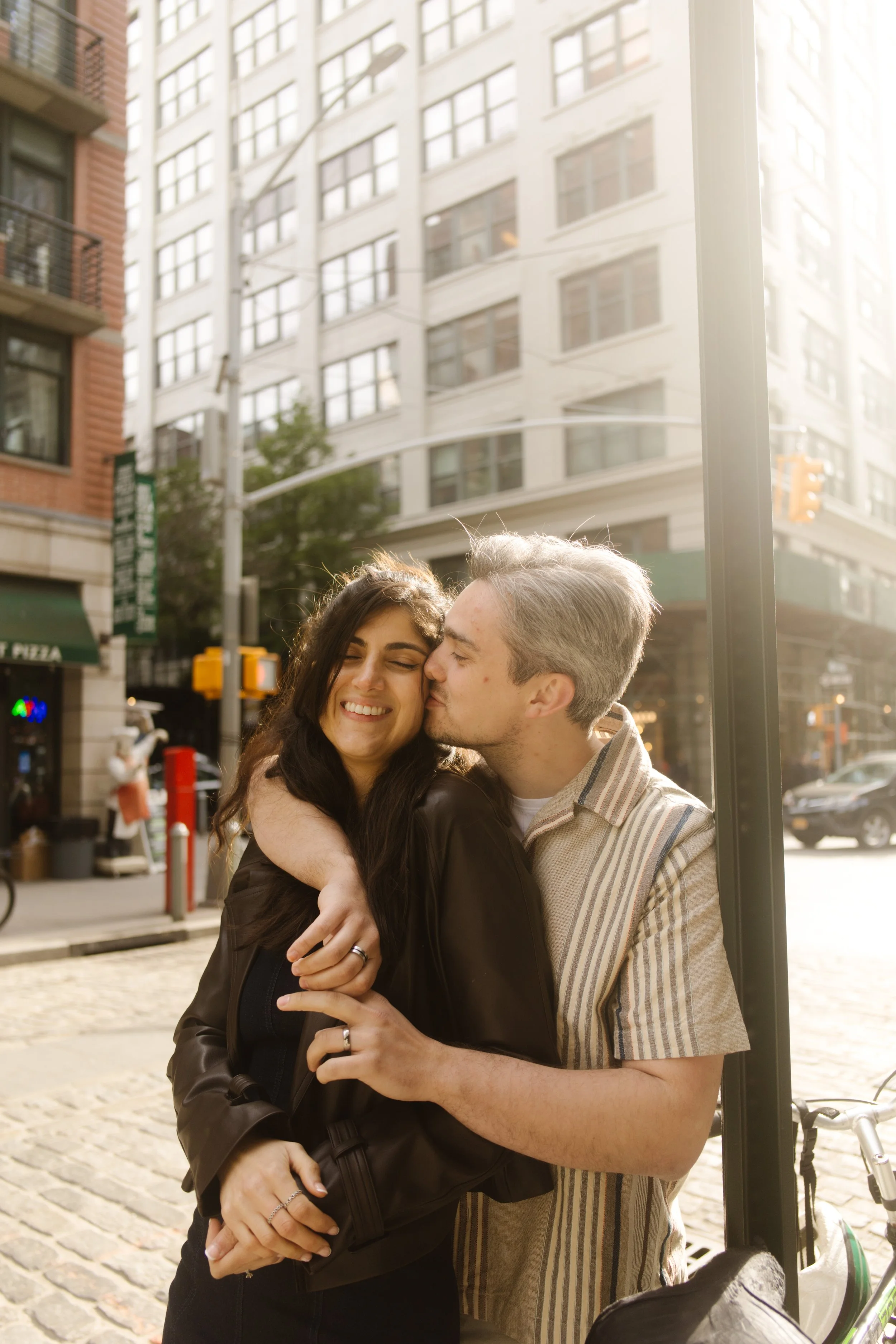 Couple embraces on a sunlit cobblestone street in DUMBO beneath the Manhattan skyline during their engagement session.