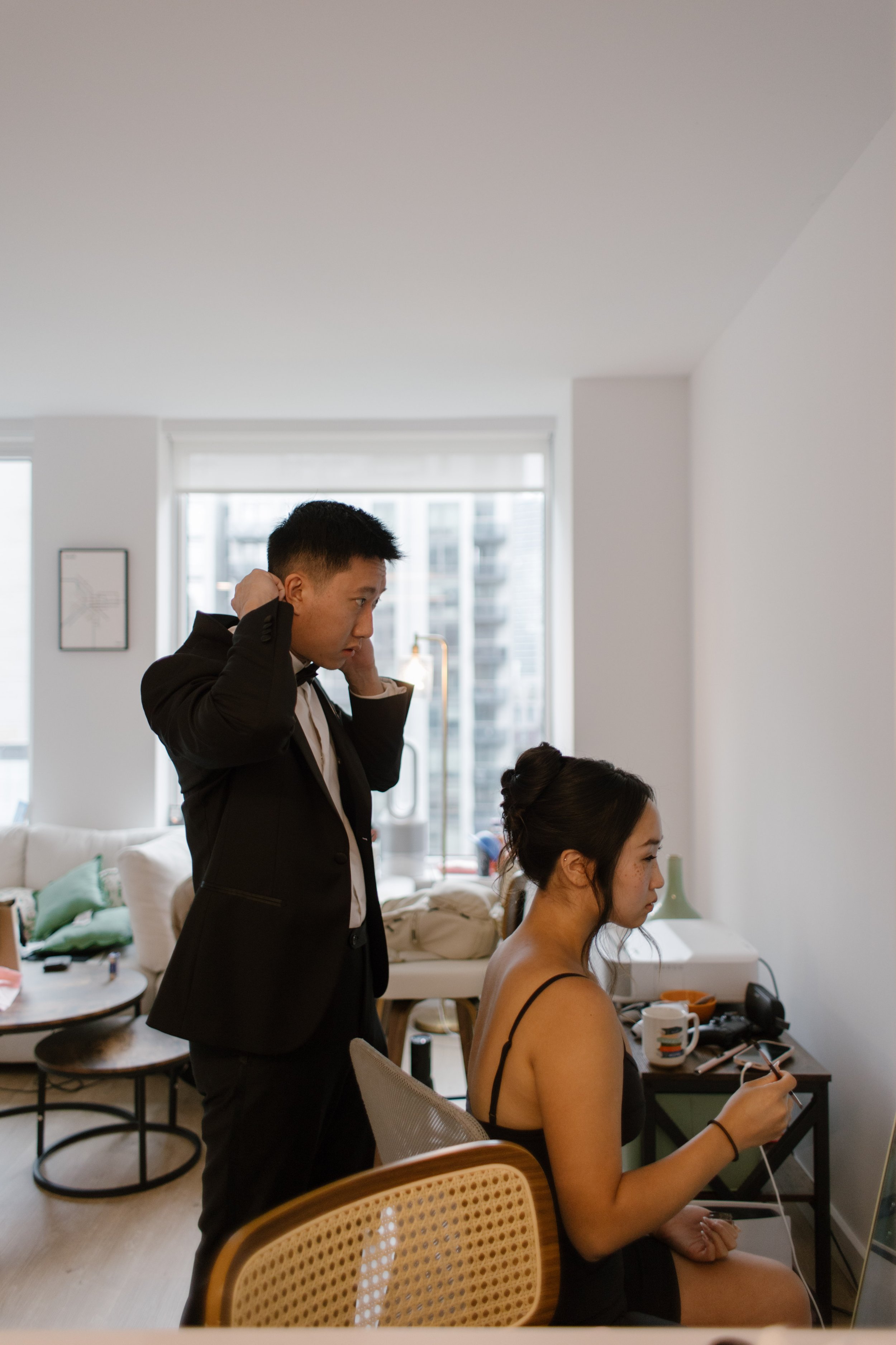 Bride getting ready while the groom adjusts his tuxedo in their apartment before heading to their NYC courthouse wedding.