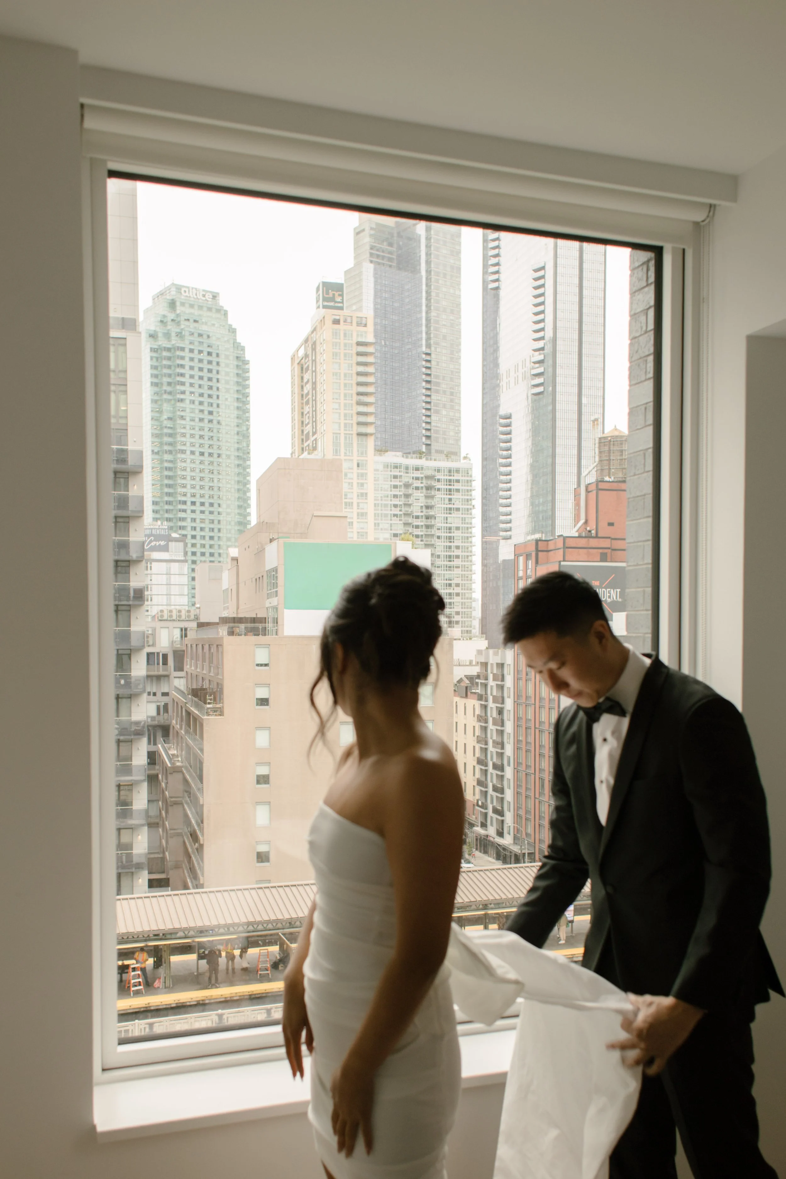 Groom helping the bride into her wedding dress in front of a large apartment window overlooking the Manhattan skyline.