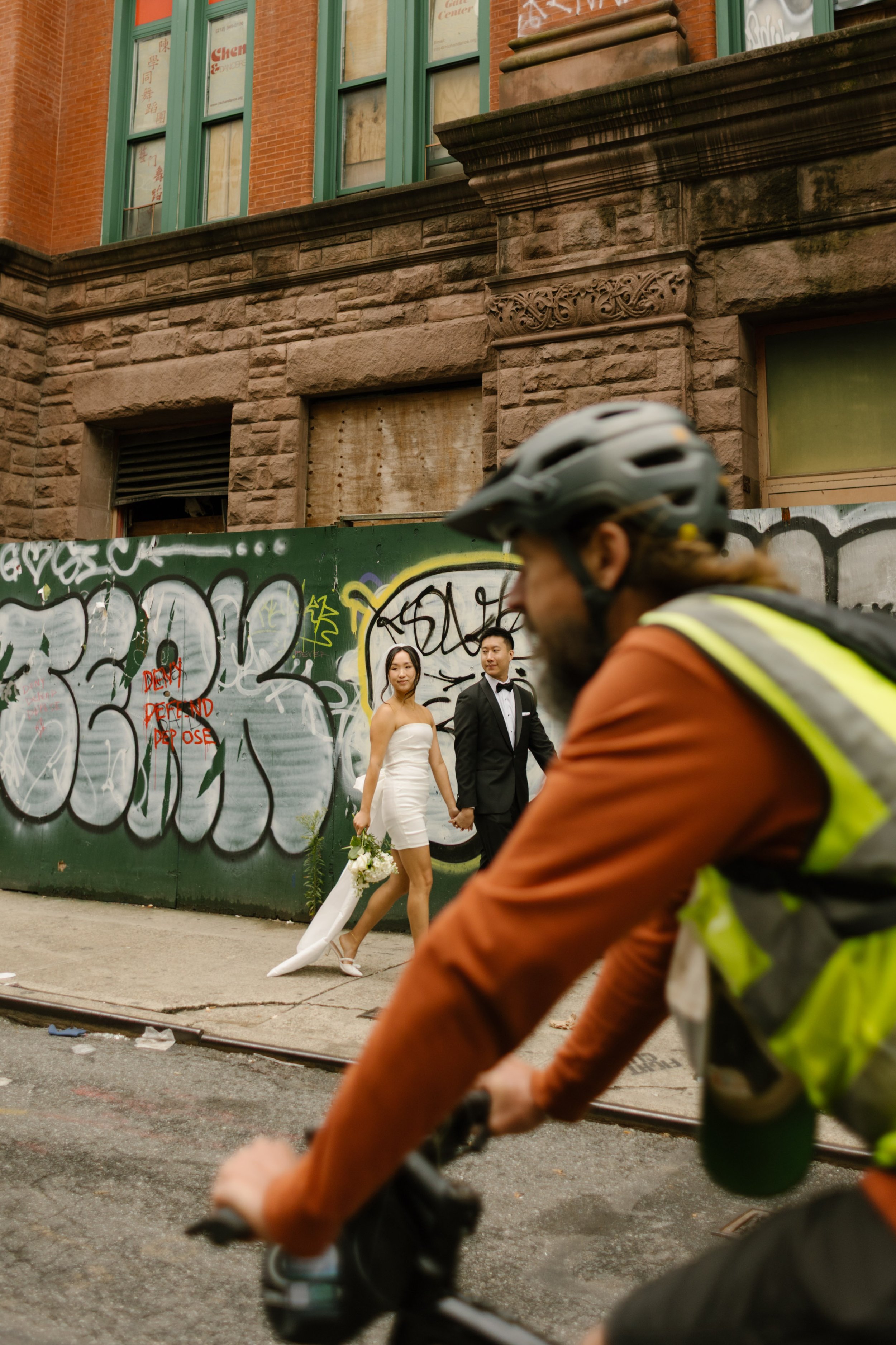 Bride and groom walking through Chinatown after their NYC courthouse wedding as a cyclist passes by, capturing an authentic New York City moment.