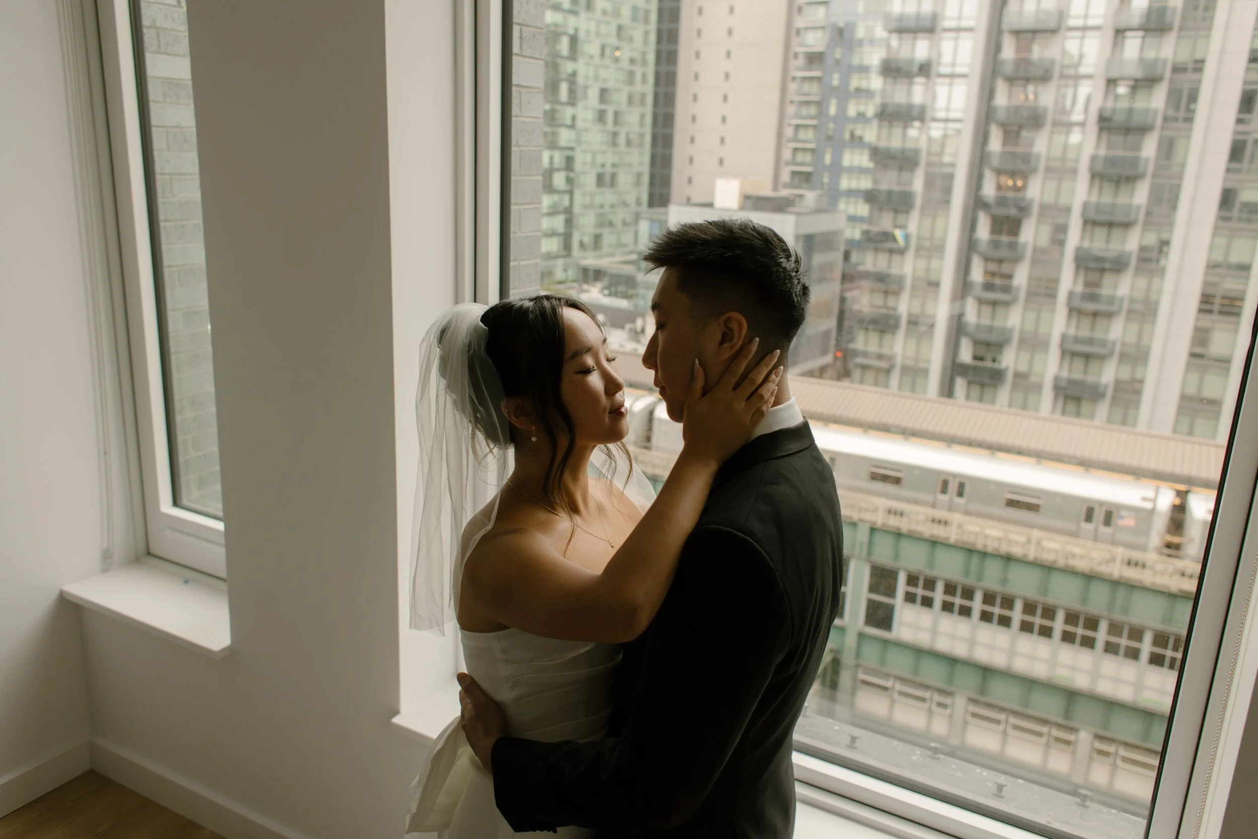 Bride and groom embracing in front of a large apartment window overlooking the Manhattan skyline before their NYC courthouse wedding.