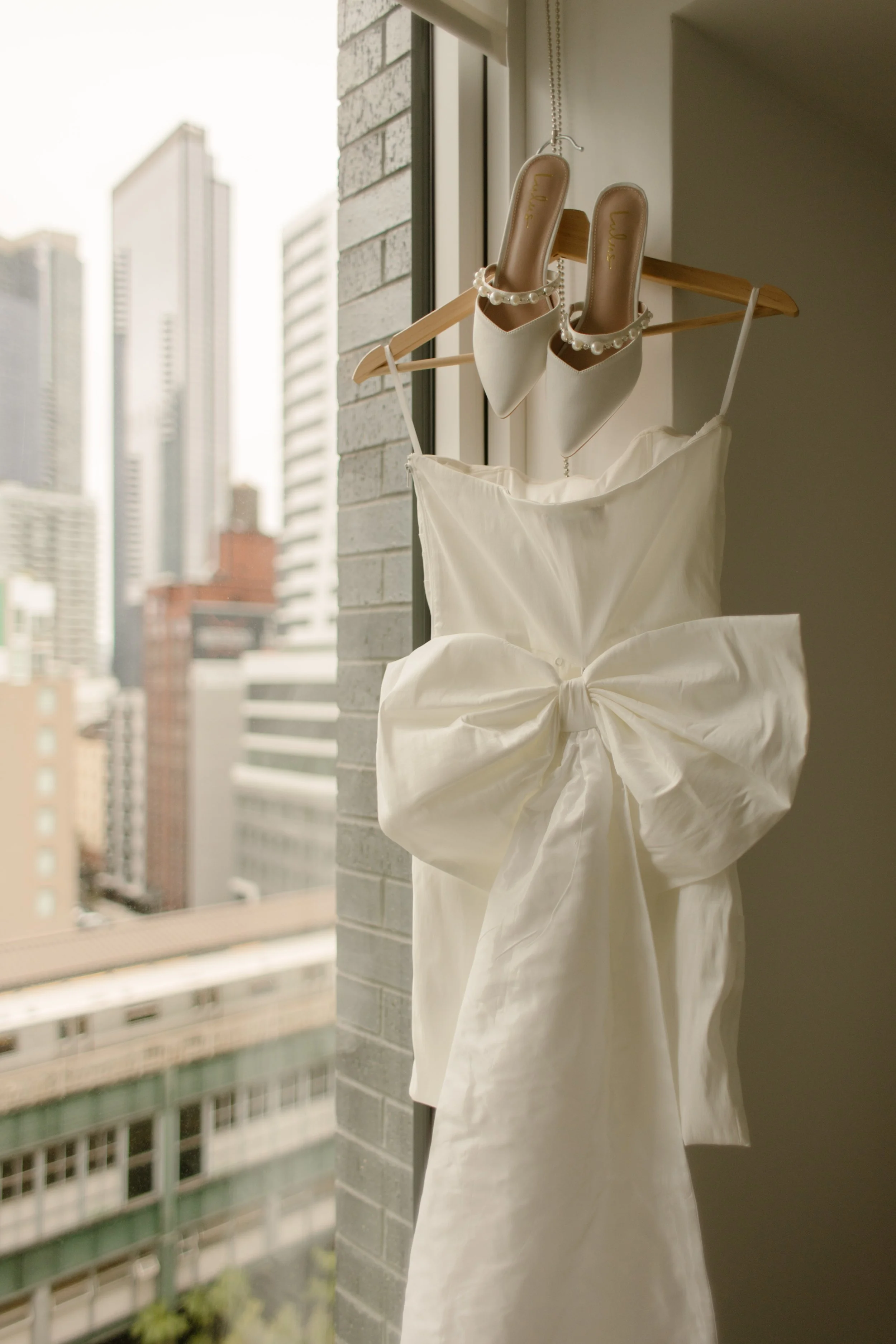 Minimal wedding dress and pearl heels hanging in an apartment window overlooking the Manhattan skyline before the ceremony.