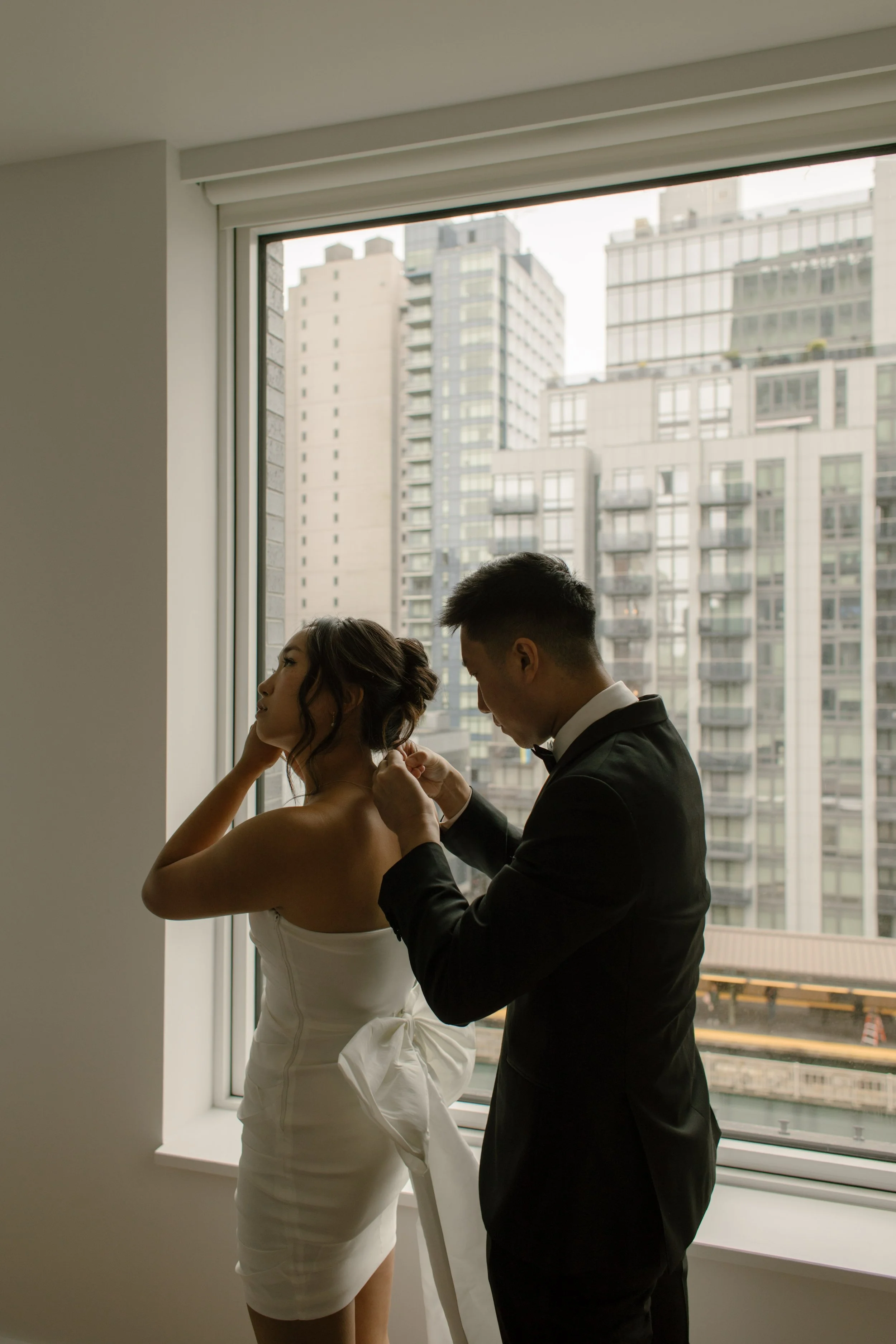 Groom fastening the bride's wedding dress in front of a large apartment window before their NYC courthouse wedding.