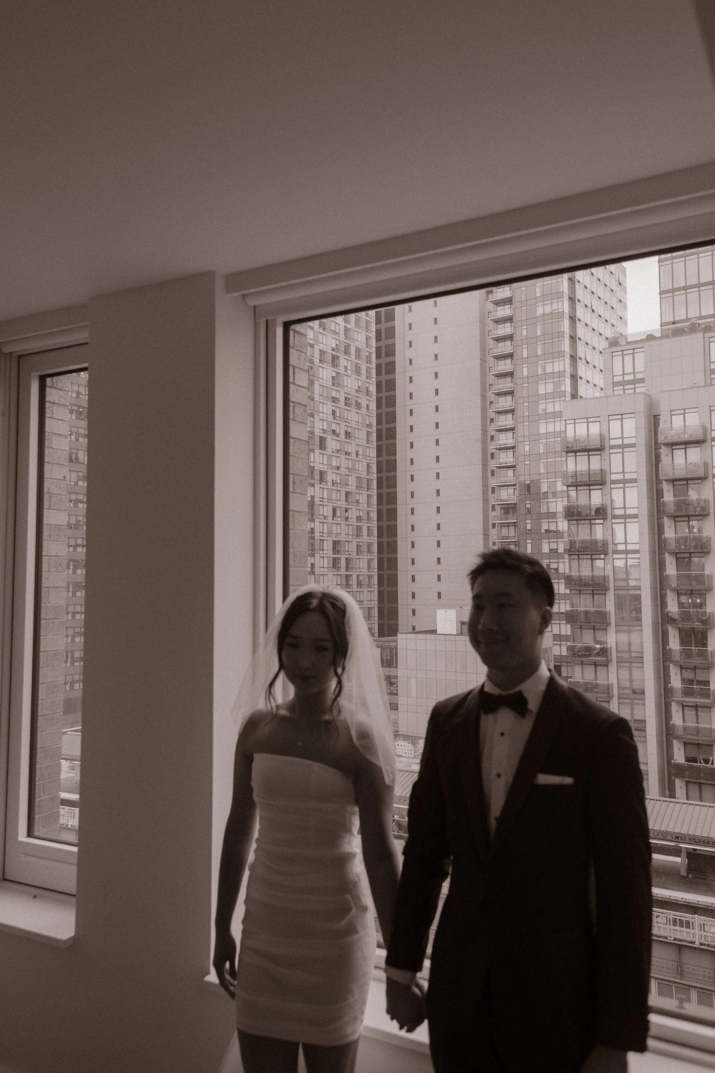 Bride and groom holding hands in front of a large apartment window before leaving for their NYC courthouse wedding.