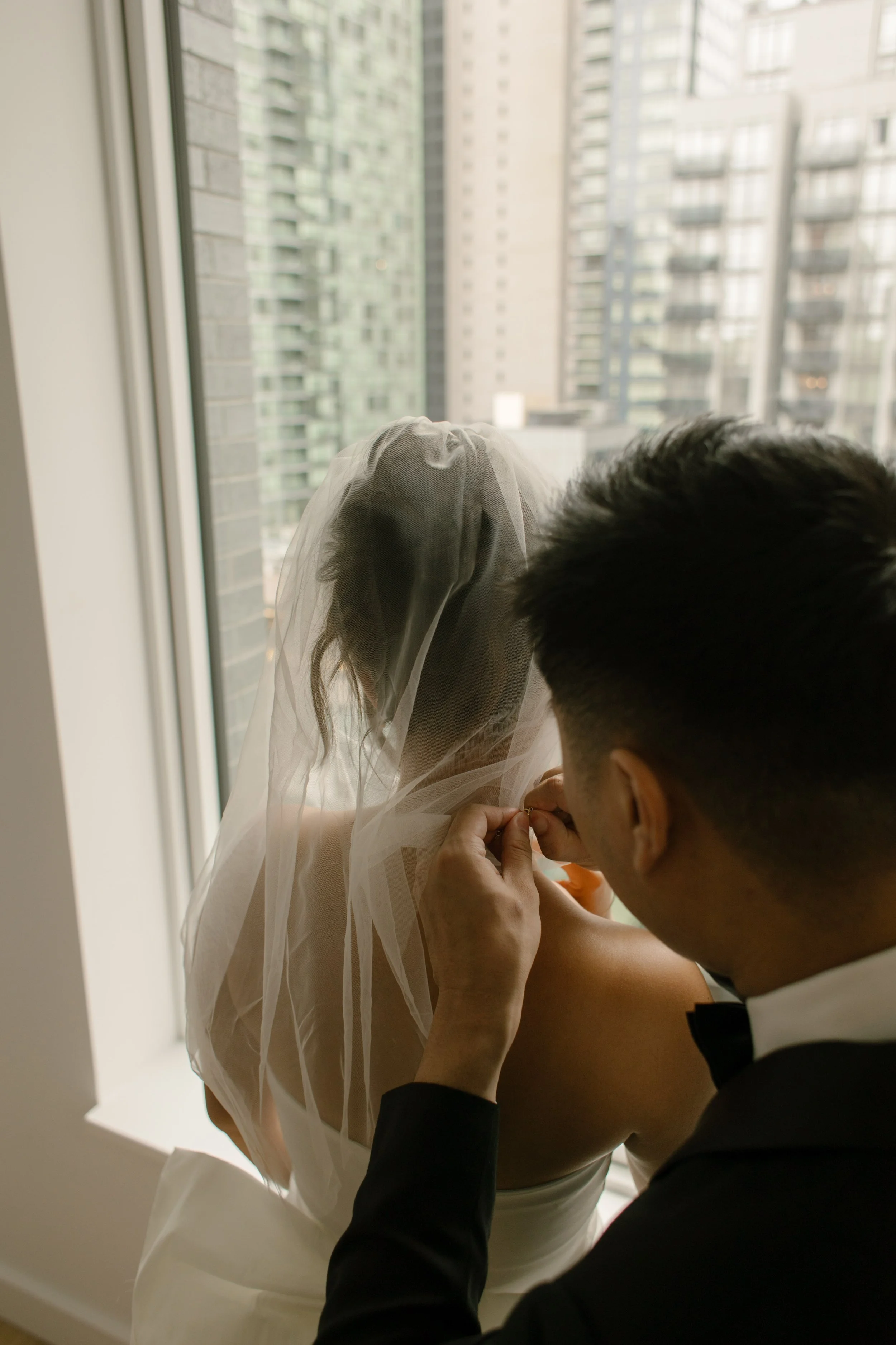 Groom fastening the bride's necklace by a large apartment window before their NYC courthouse wedding.