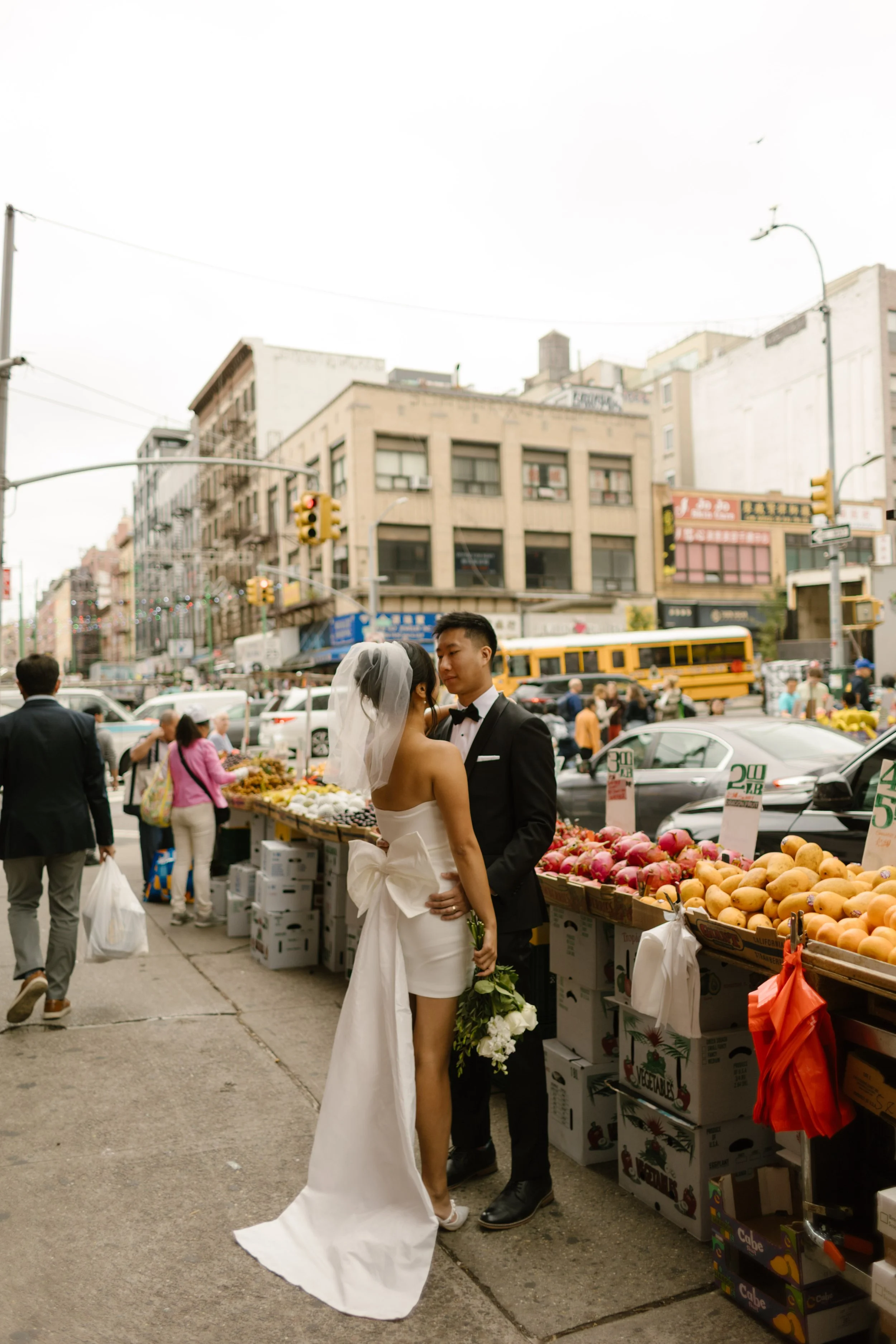 Bride and groom embrace beside Chinatown fruit stands during portraits after their New York City courthouse ceremony.