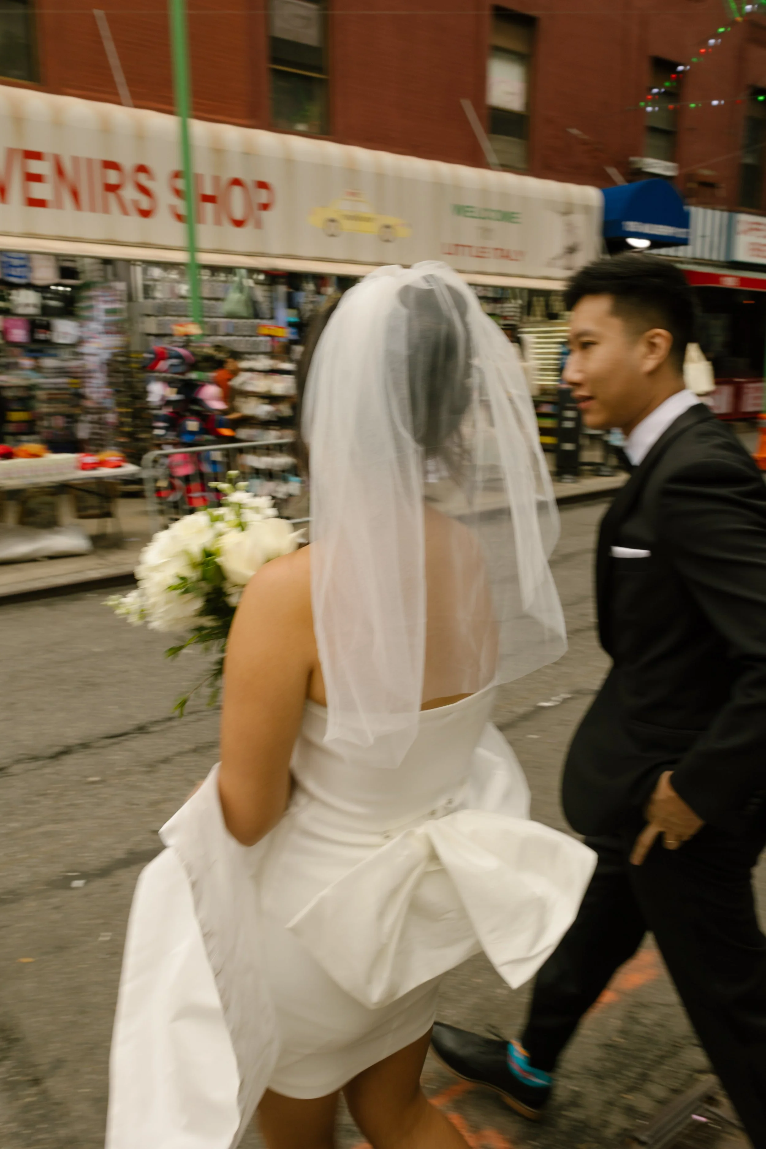 Bride and groom walk through a lively New York City street lined with neighborhood shops following their courthouse wedding celebration.