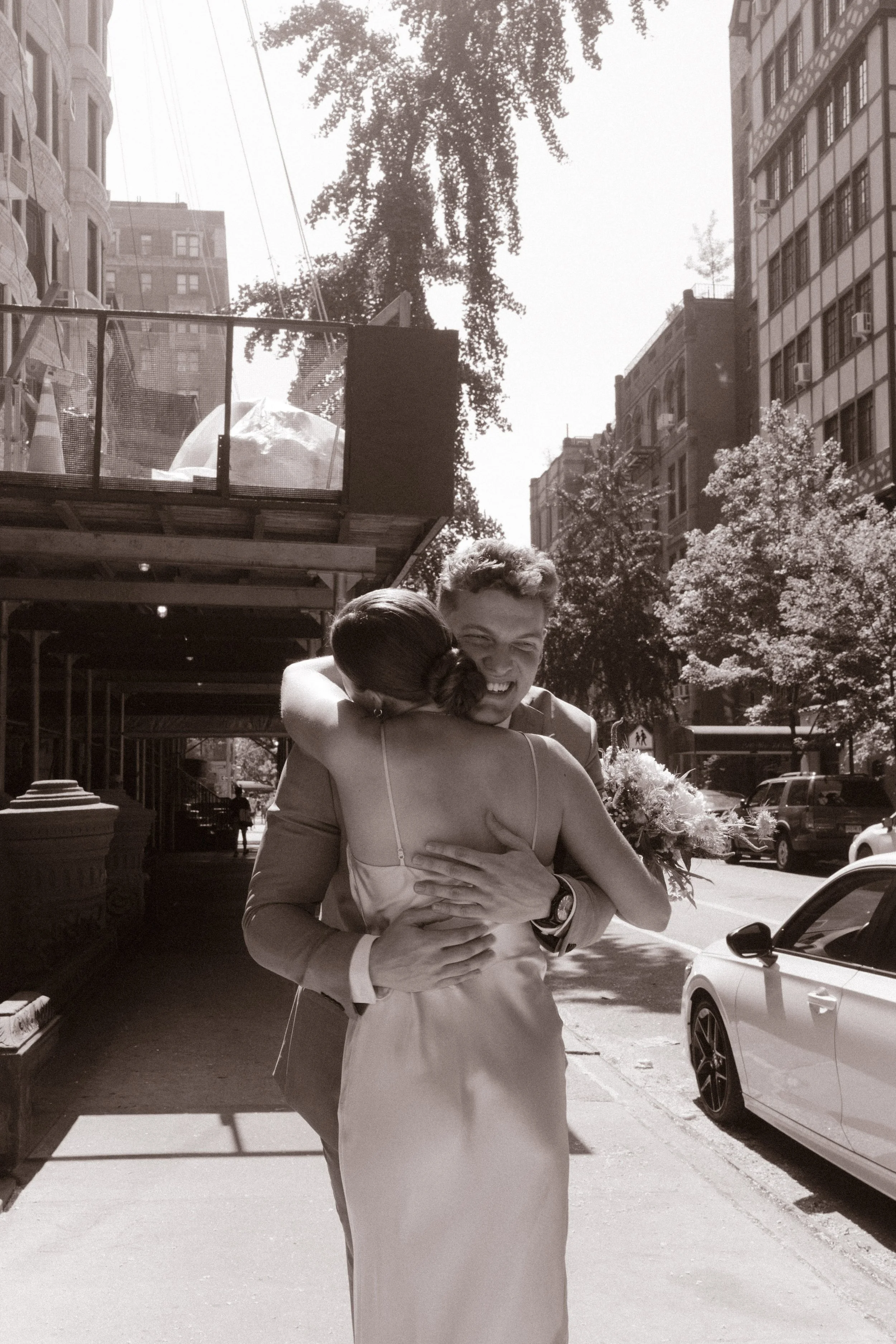Black-and-white photo of newlyweds embracing on a sunlit Manhattan street.