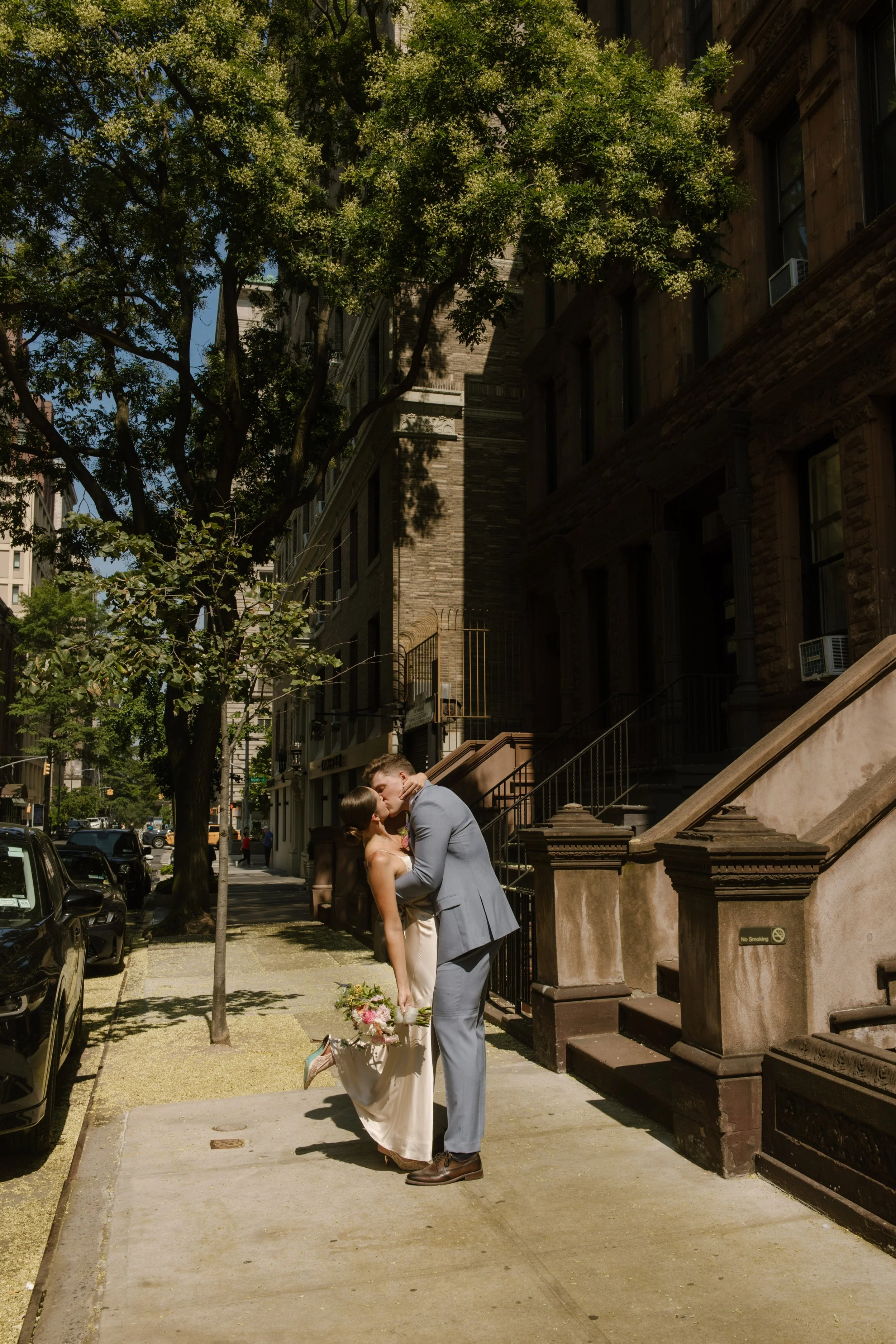 Bride and groom sharing a kiss on a quiet brownstone-lined street during a pause in their wedding day timeline.