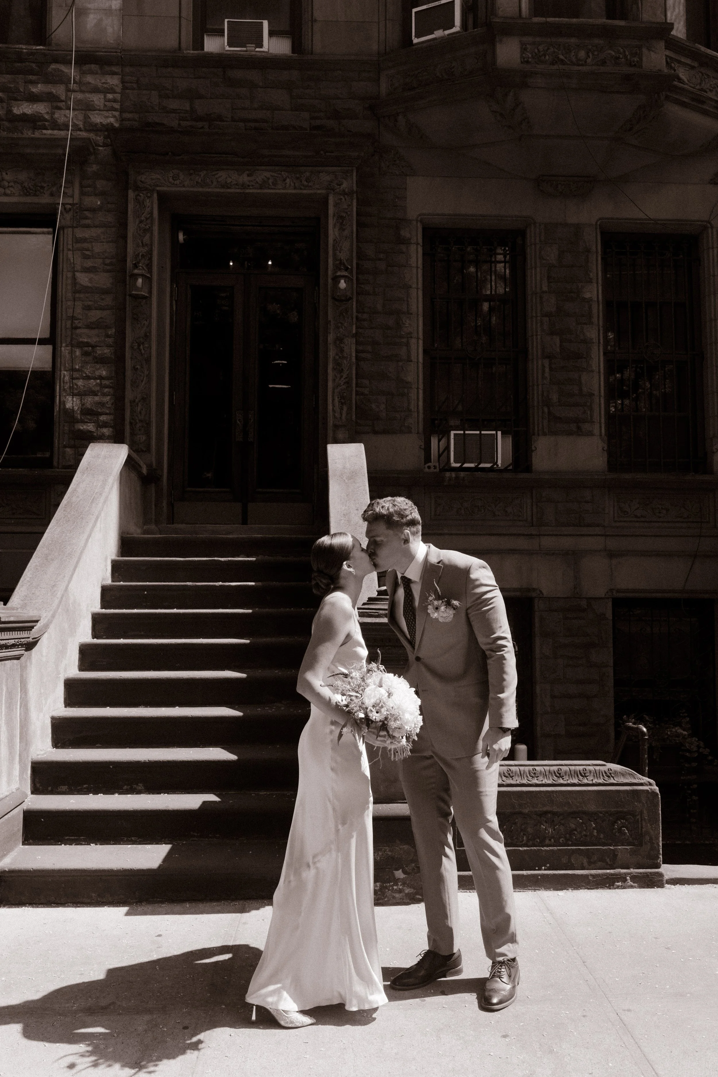 Black-and-white portrait of the bride and groom kissing outside a classic New York brownstone during their wedding day timeline.