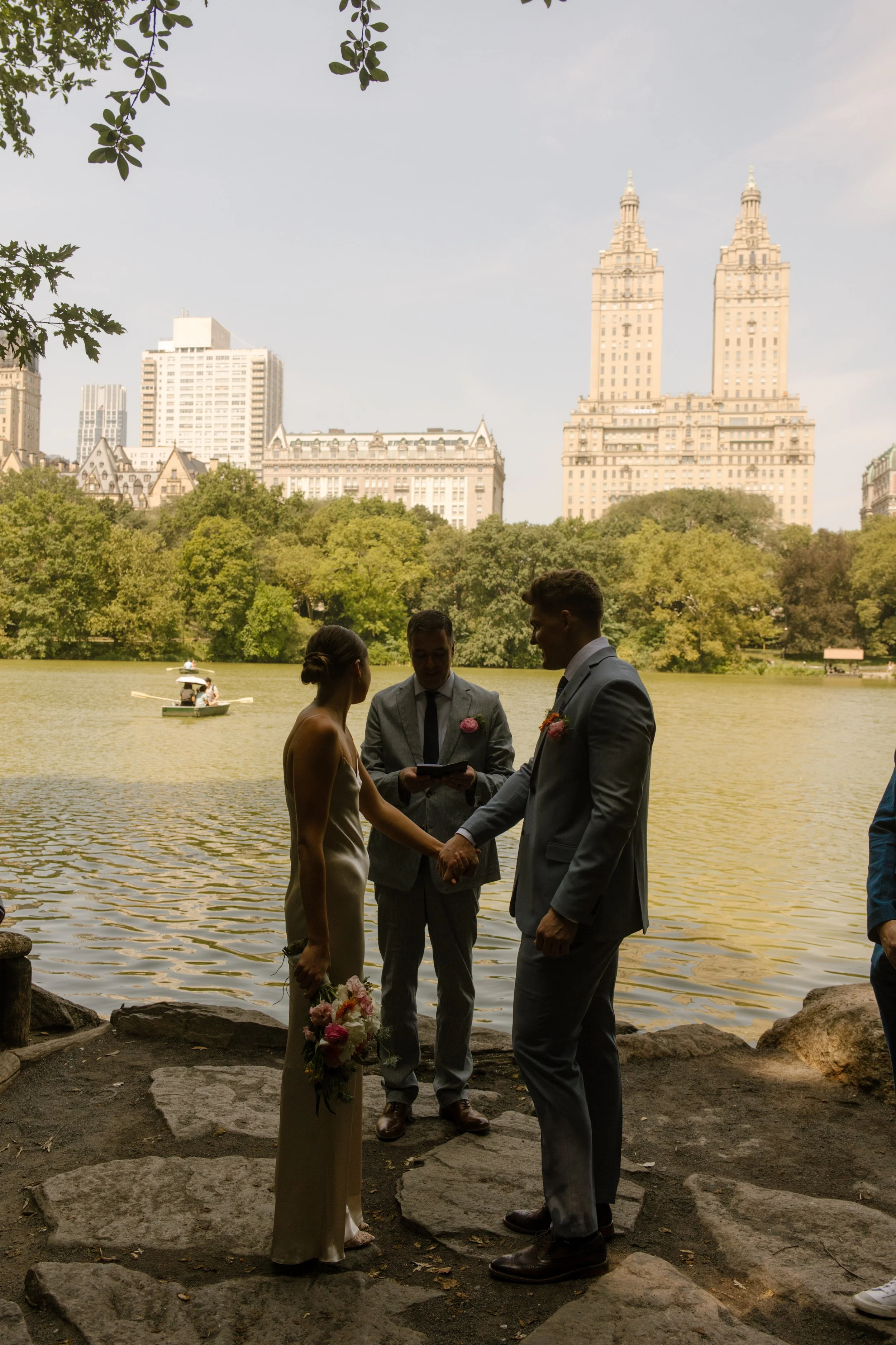 Bride and groom holding hands during their lakeside ceremony with the New York City skyline behind them as part of their wedding day timeline.