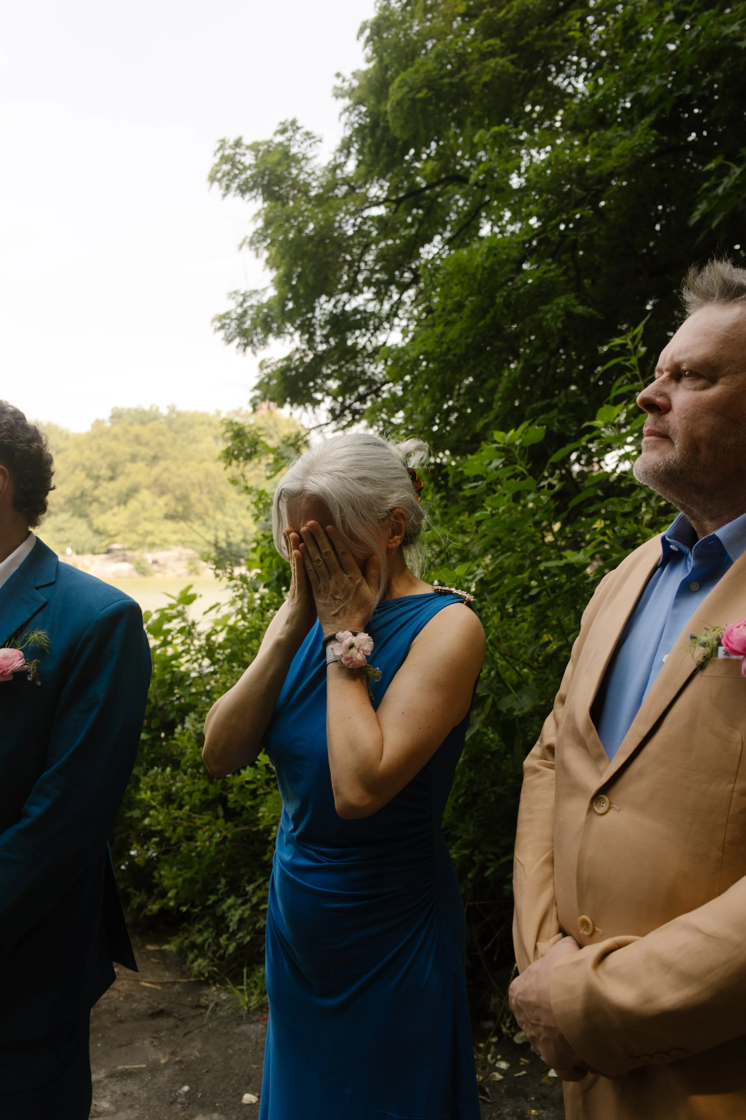 Emotional mother of the groom wiping away tears during an outdoor lakeside ceremony, a heartfelt moment in the wedding day timeline.