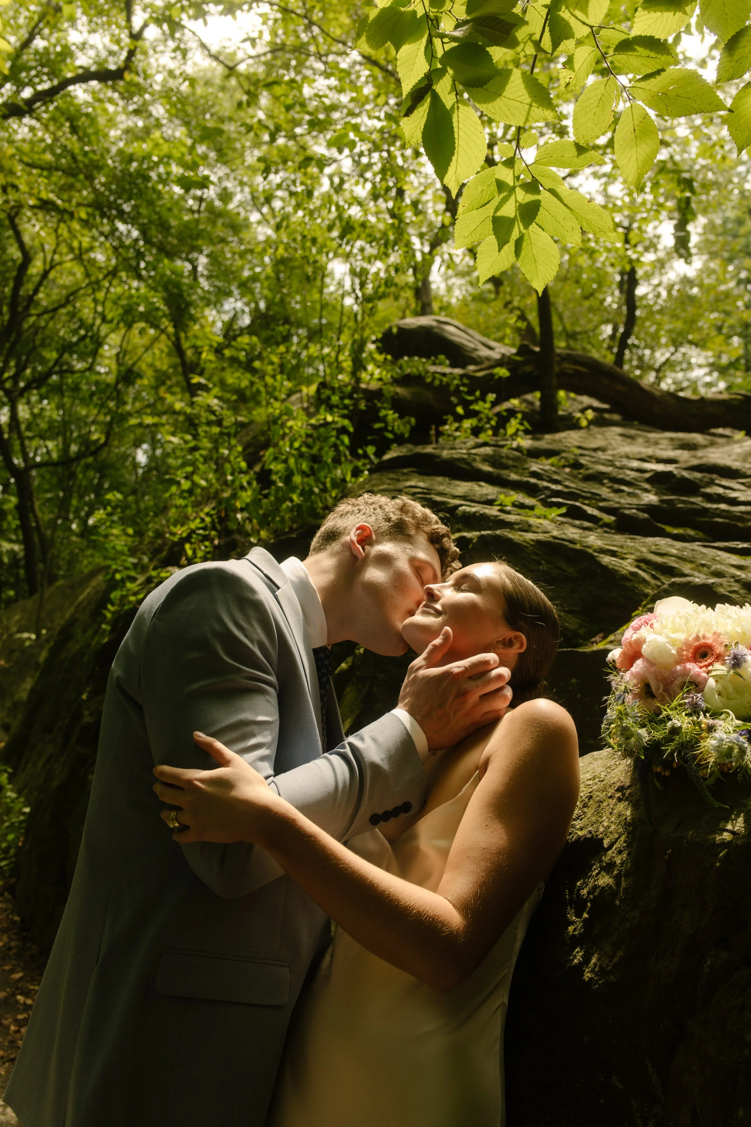 Bride and groom sharing a kiss beneath leafy trees after their ceremony during their wedding day timeline.