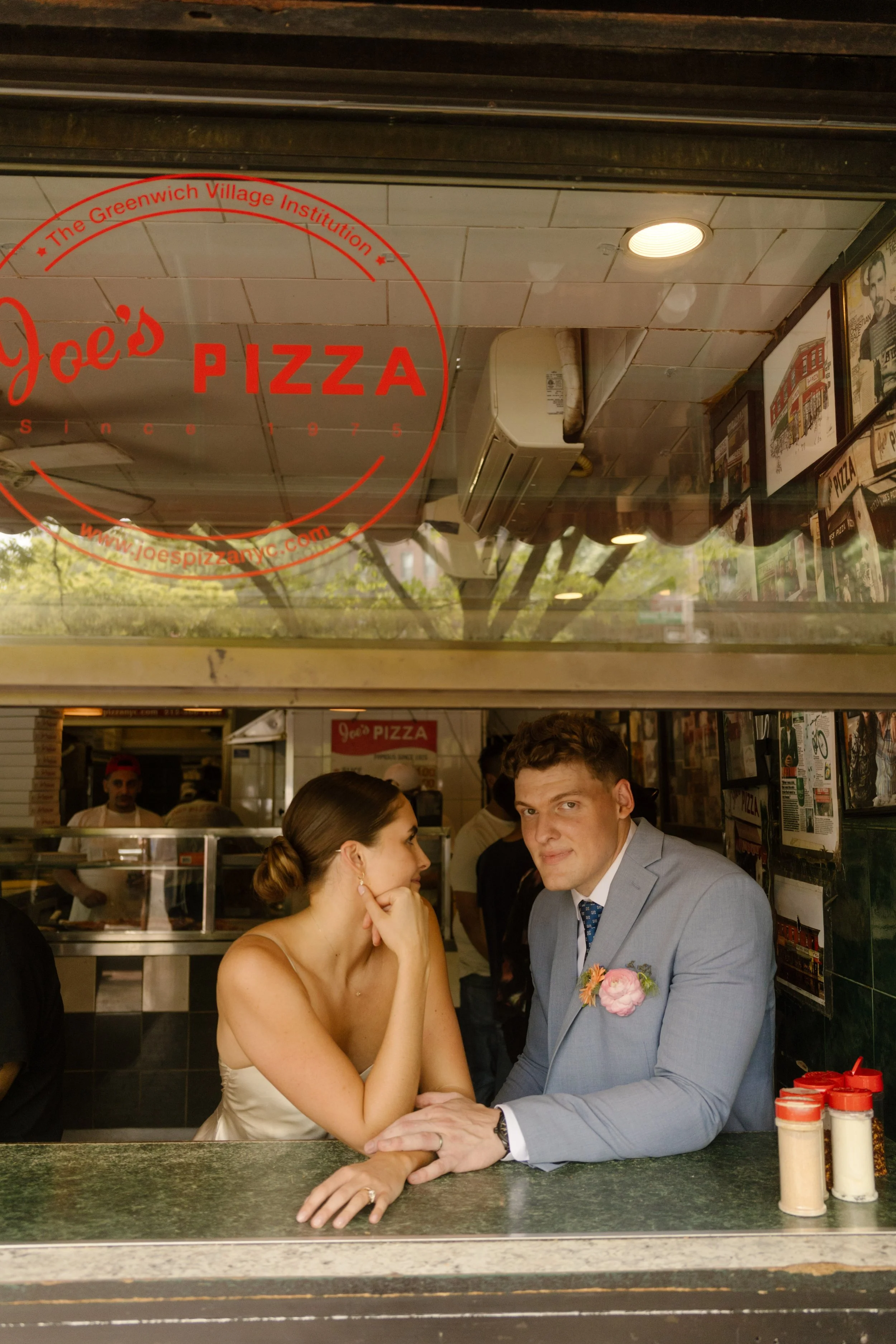 Bride and groom sitting together inside a classic New York pizza shop during a relaxed break in their wedding day timeline.