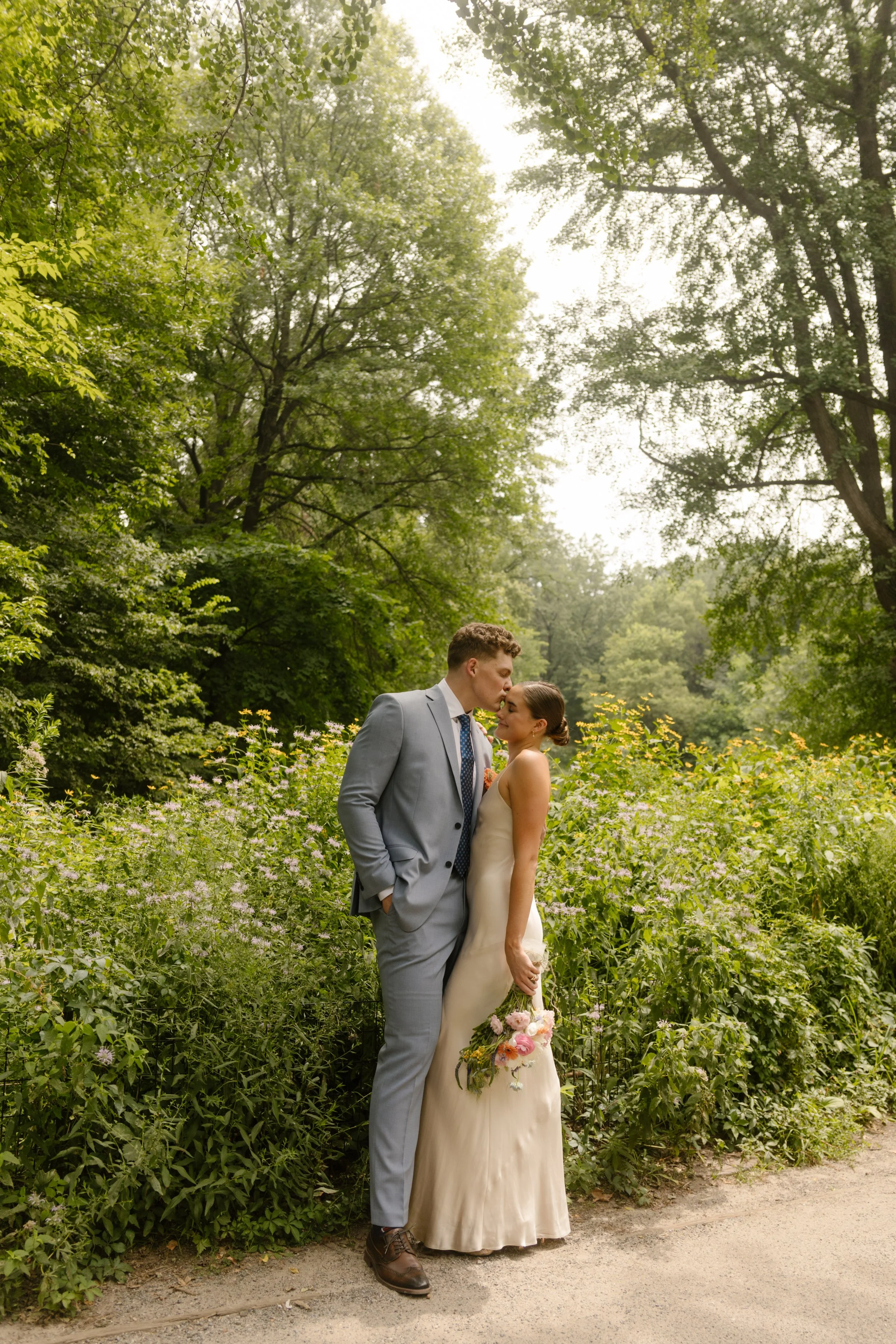 Newlyweds sharing a kiss along a garden path surrounded by summer greenery during their wedding day timeline.
