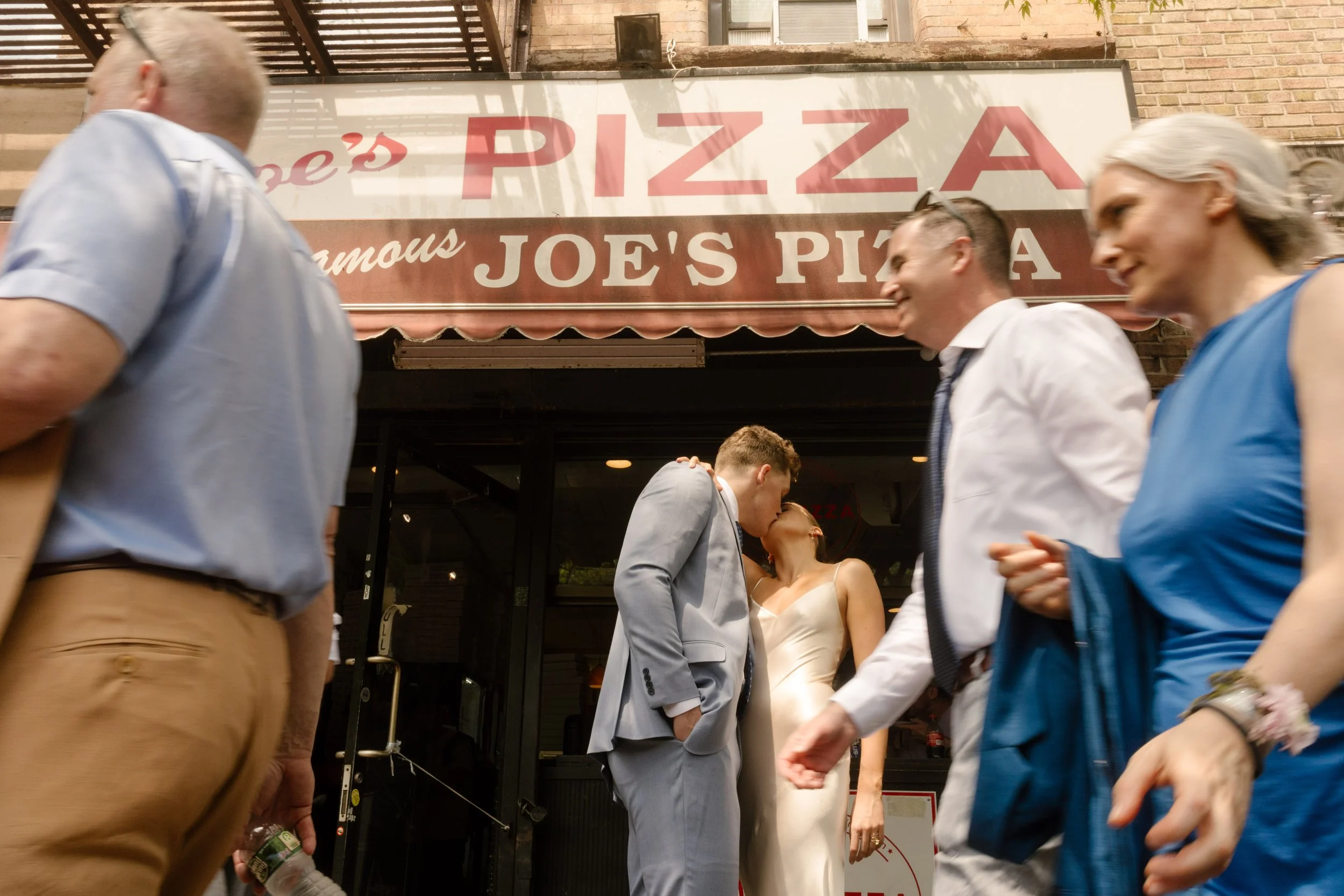 Couple kissing outside Joe's Pizza as pedestrians walk by on a busy Manhattan street.