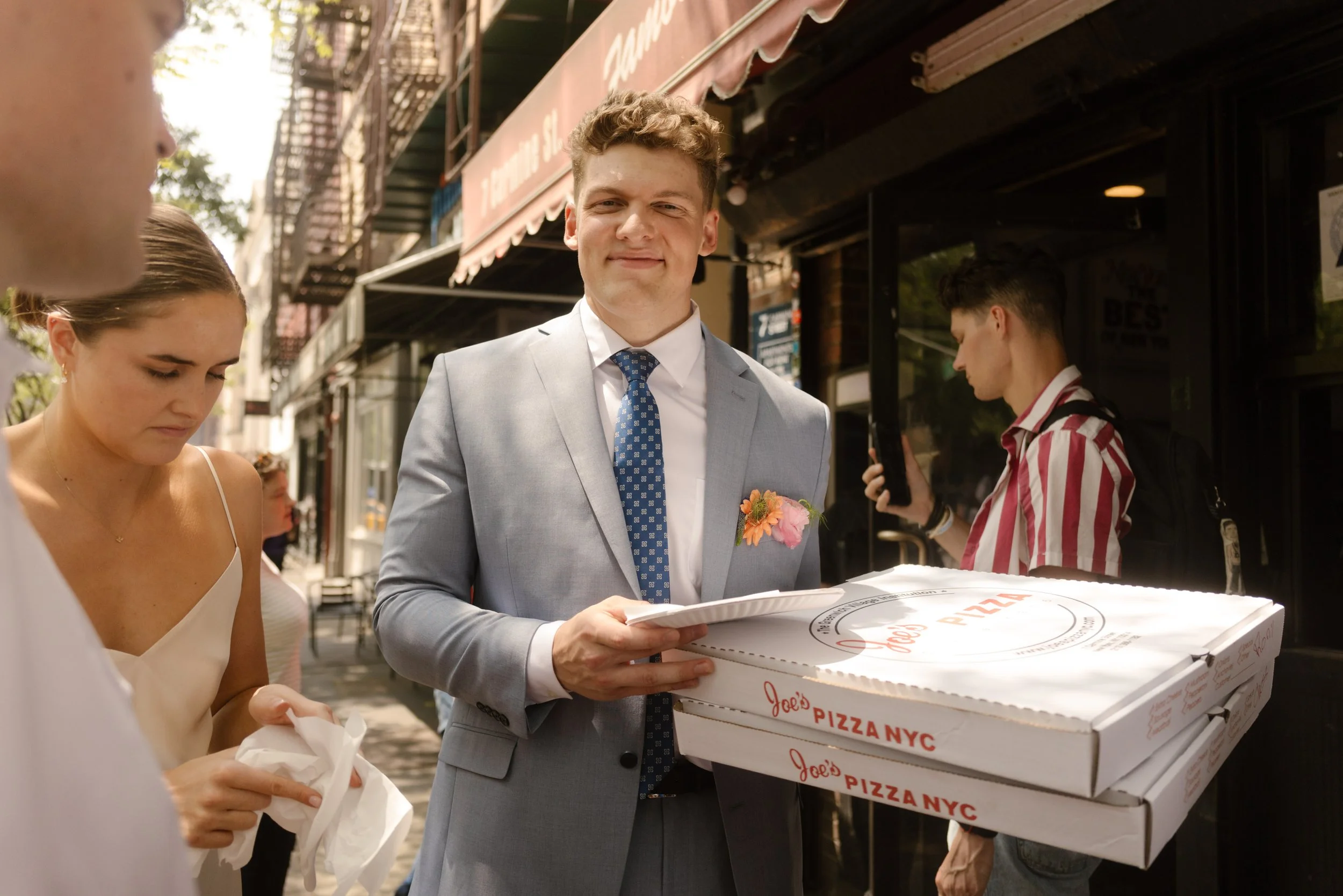 Groom carrying fresh pizza boxes outside a New York pizzeria during a fun stop on their wedding day timeline.