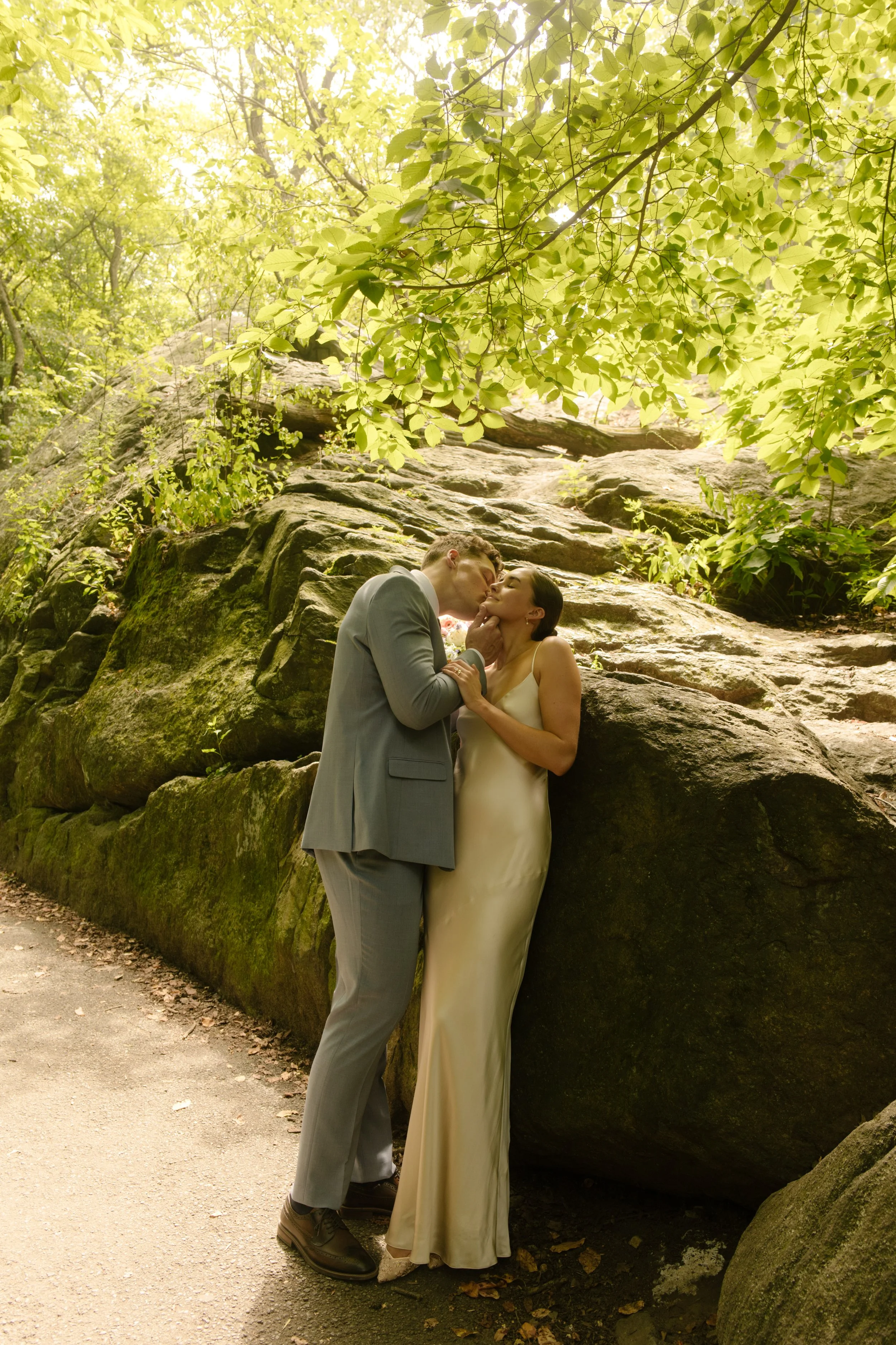 Newlyweds sharing a kiss beside large boulders in Central Park during a quiet moment in their wedding day timeline.