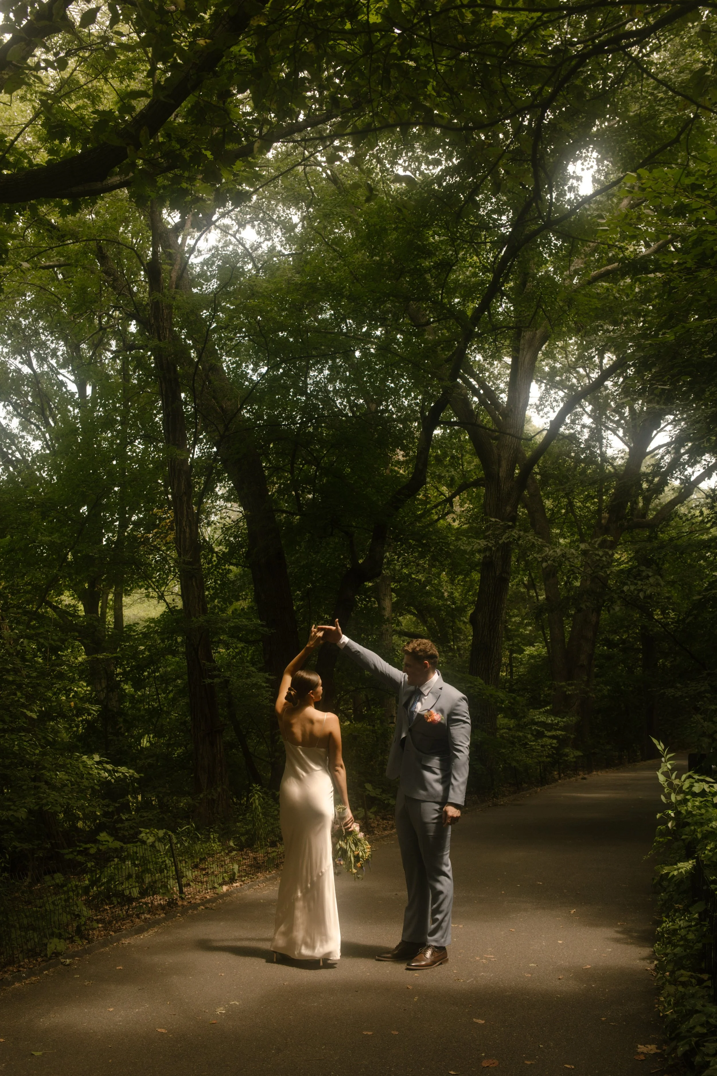 Groom twirling the bride beneath a canopy of trees during their wedding day timeline portraits.