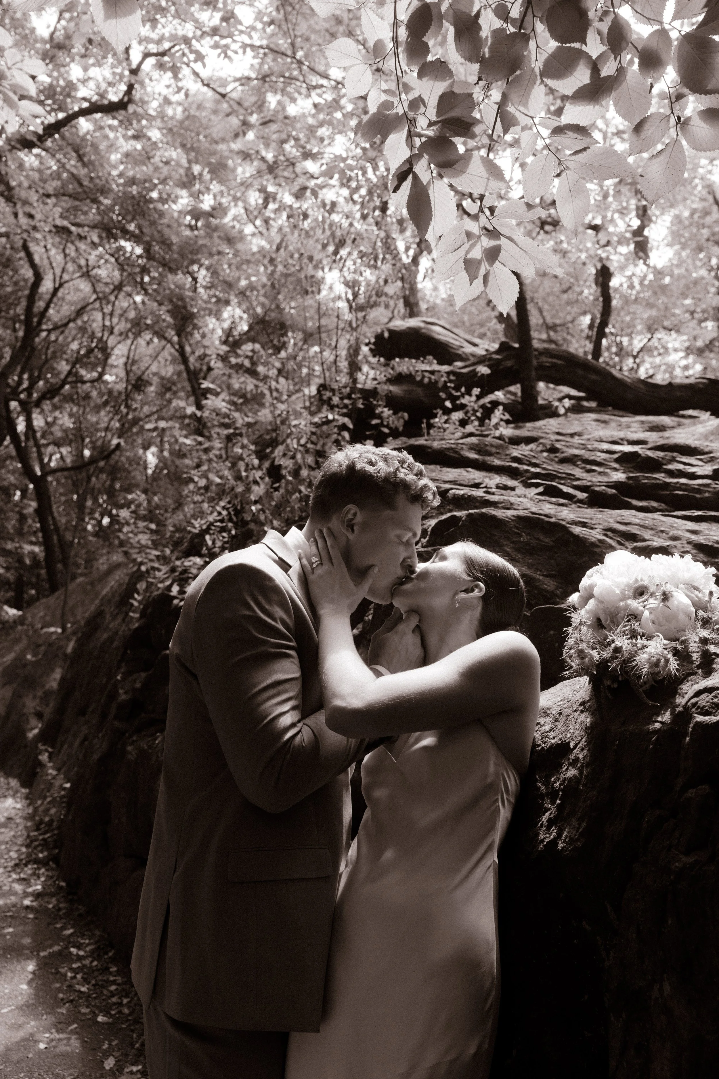 Black-and-white portrait of the bride and groom sharing a kiss beneath leafy trees in Central Park.
