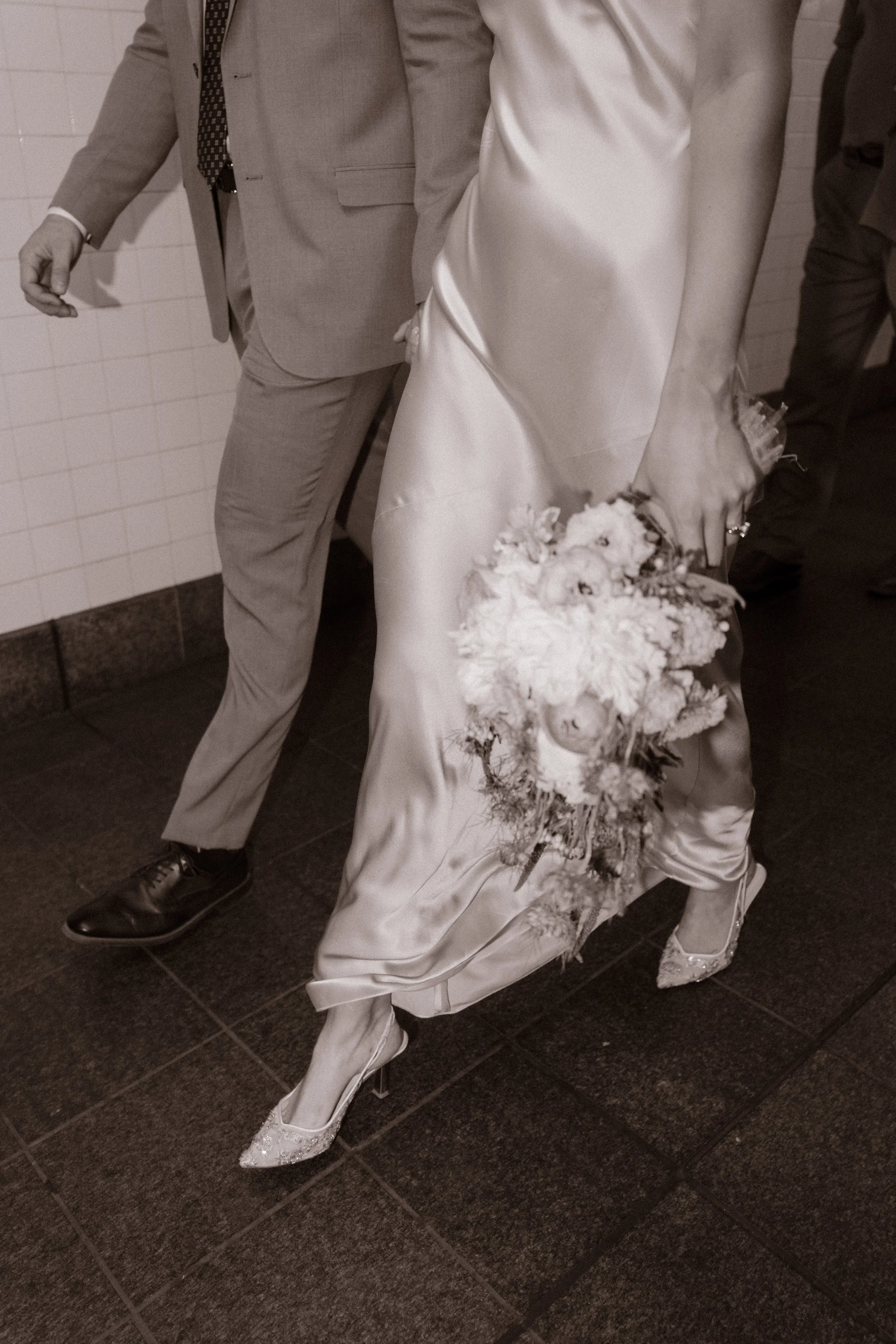 Black-and-white close-up of the bride's bouquet and wedding shoes while walking through a New York City subway station.