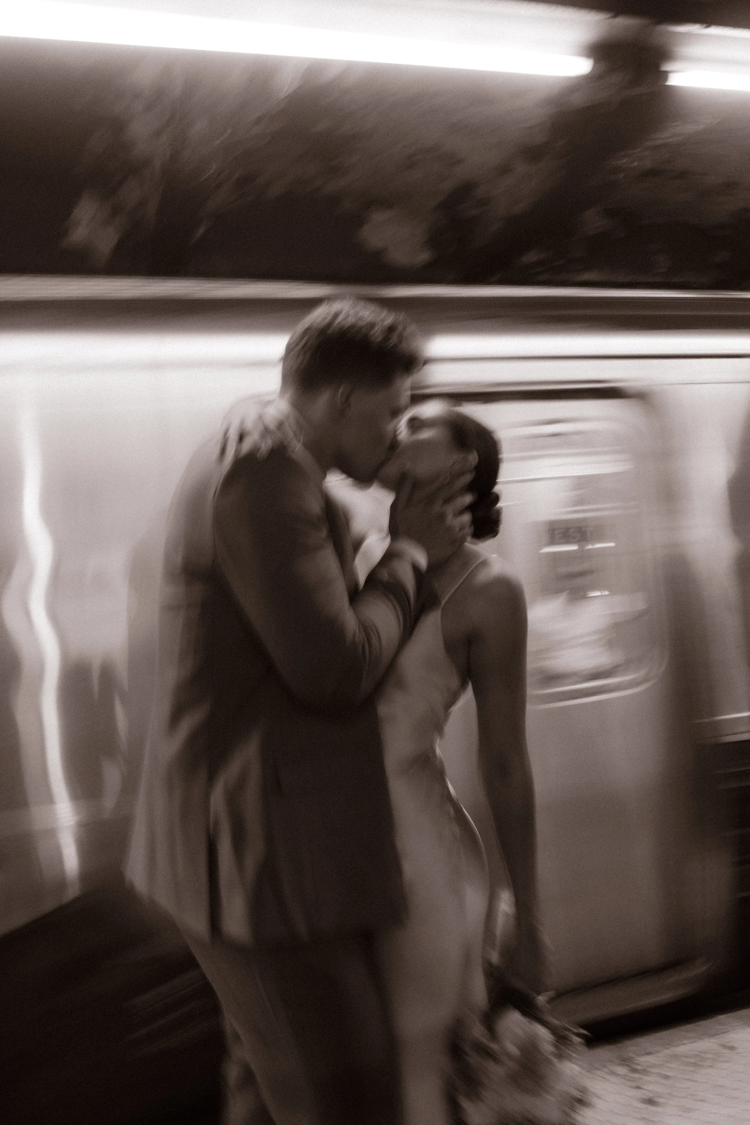 Black-and-white motion blur portrait of the newlyweds kissing on a subway platform during their wedding day timeline.