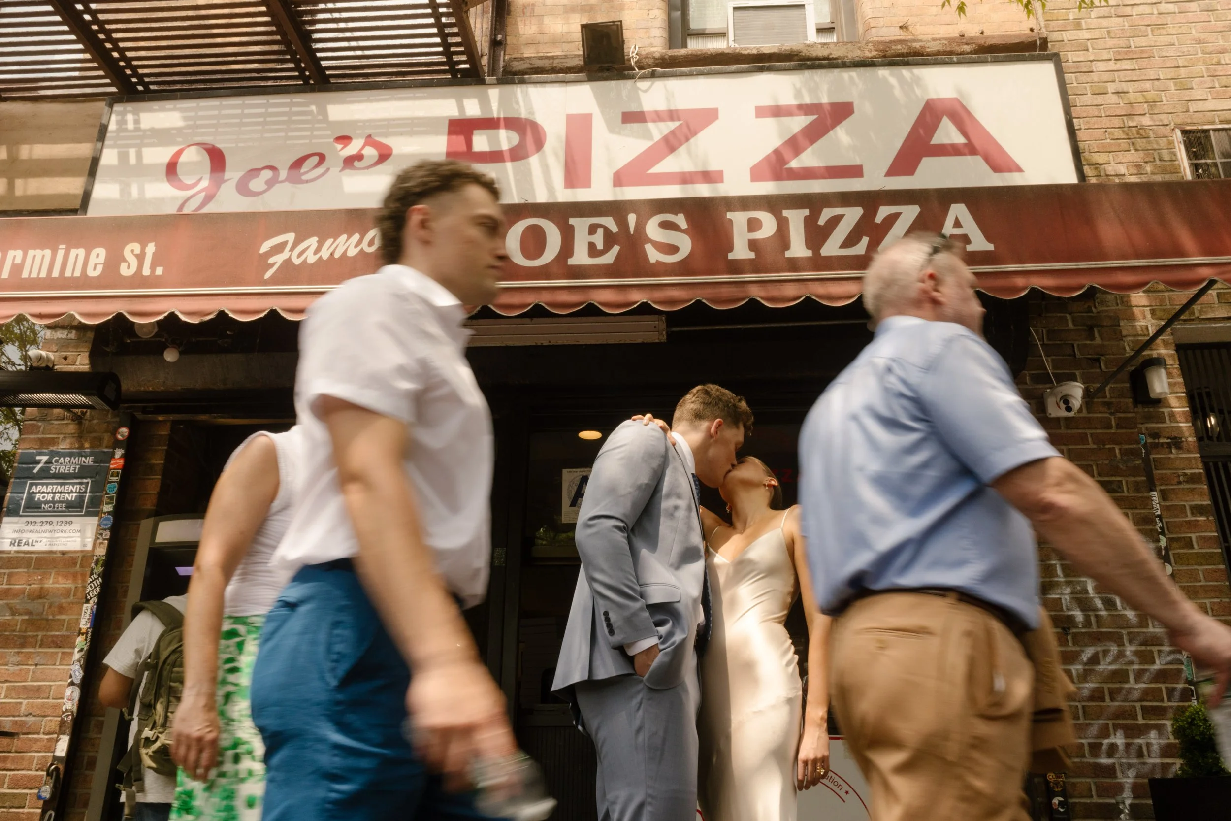Bride and groom sharing a kiss outside Joe's Pizza while pedestrians pass by, a fun stop in their wedding day timeline.