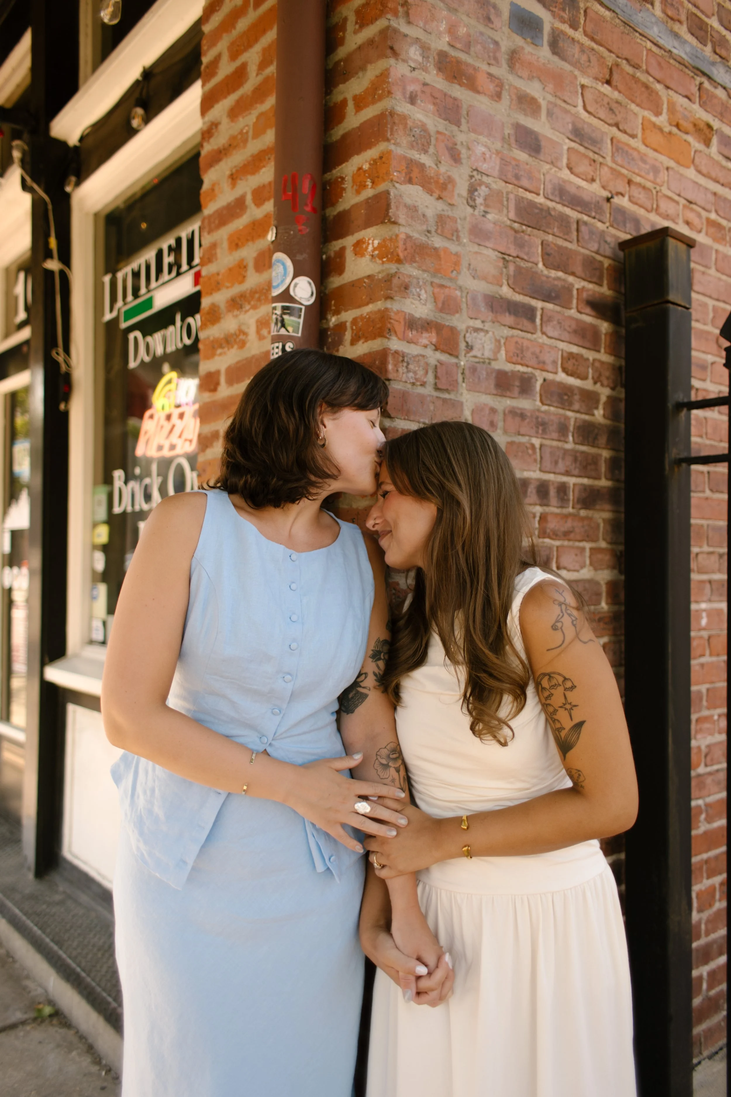 A bride kisses her partner's forehead while embracing outside a brick storefront, captured by a documentary wedding photographer.