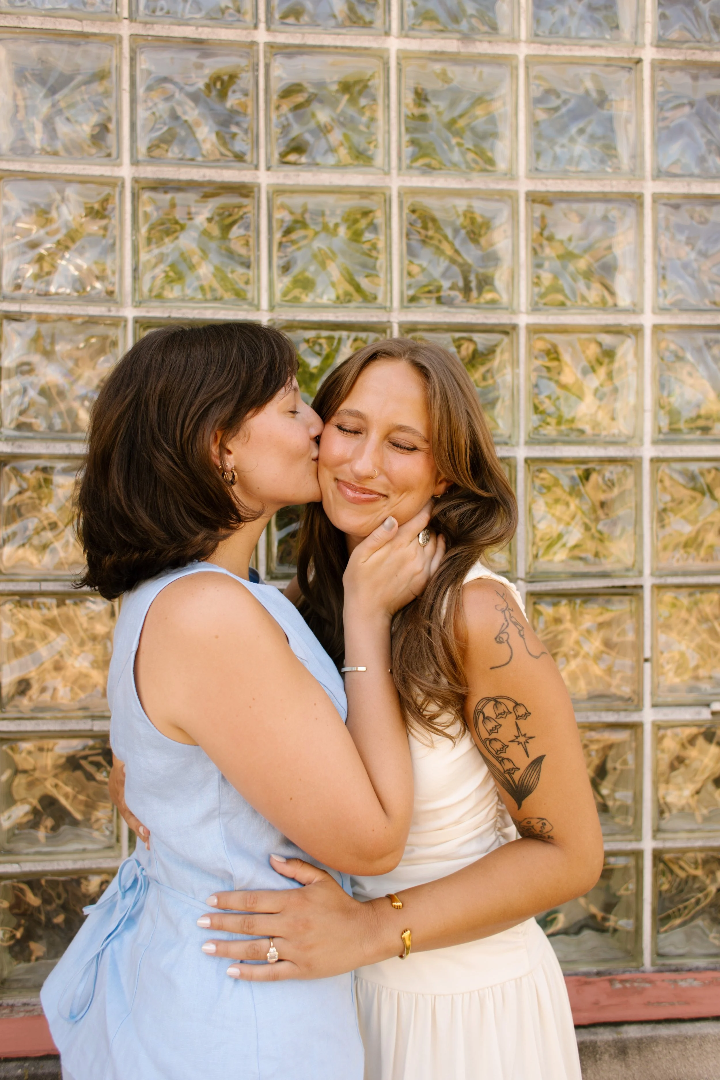 One bride kisses her partner's cheek in front of a glass block wall during intimate wedding portraits.