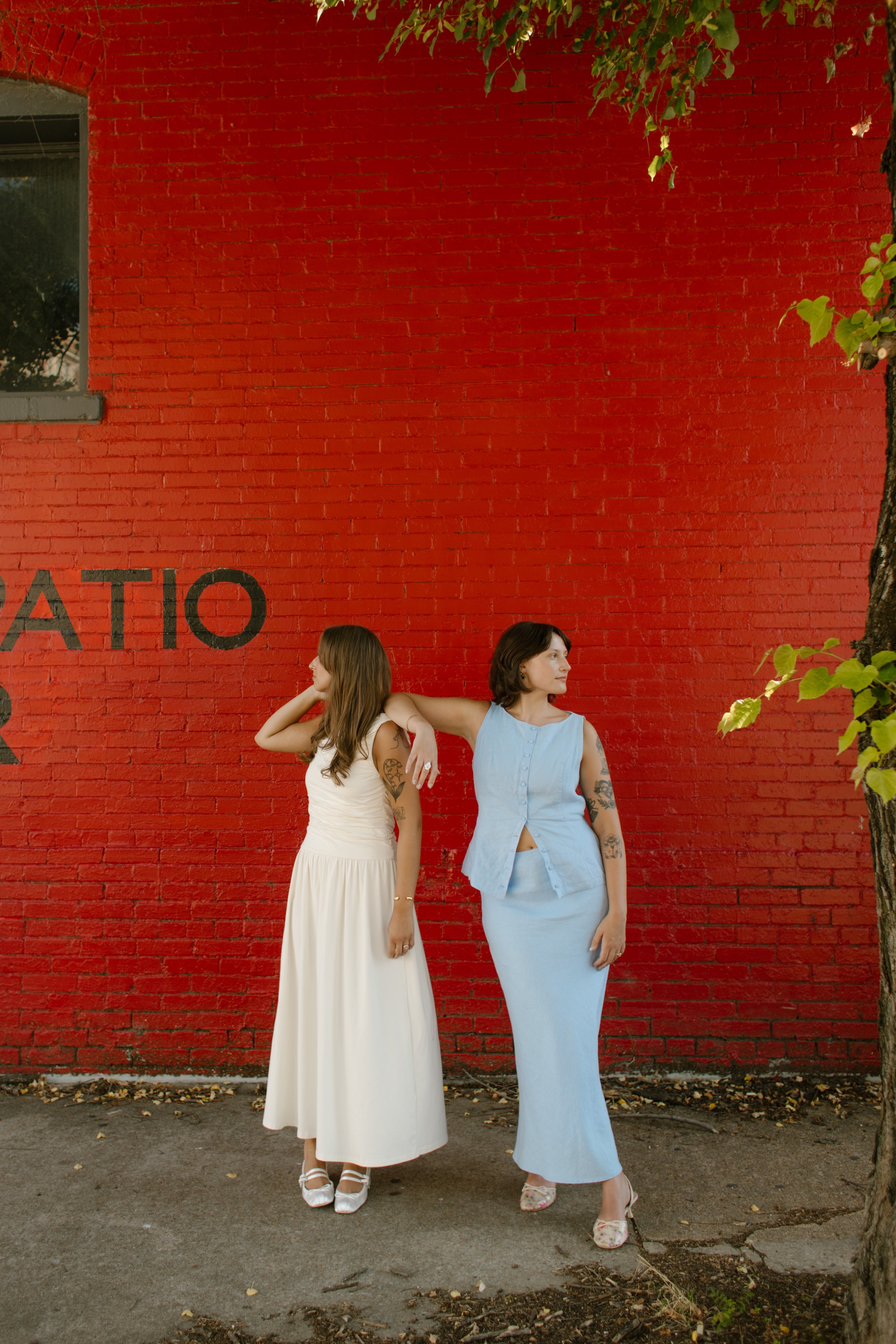 Two brides pose against a bold red brick wall with contrasting pastel dresses.