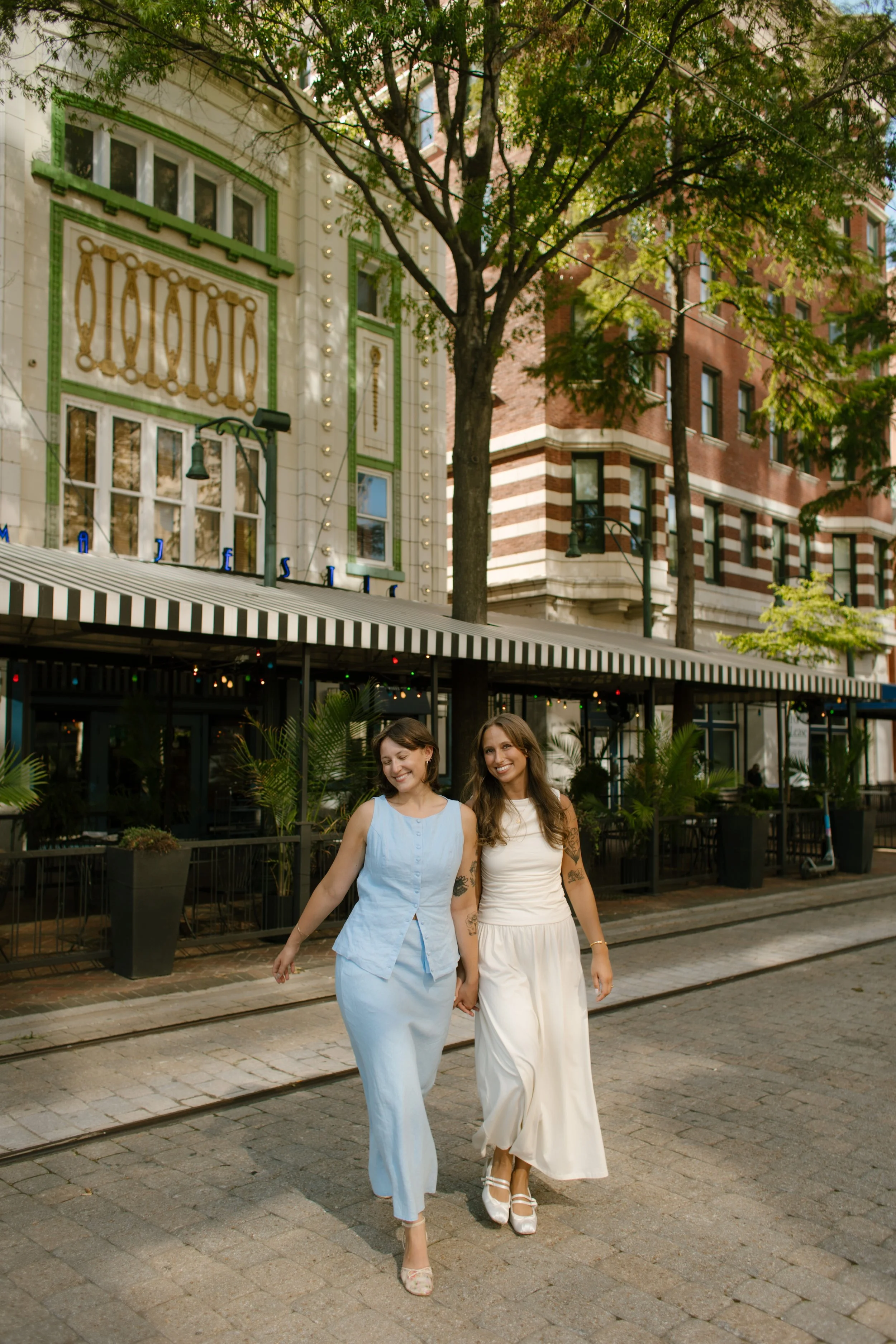 Newlyweds walk hand in hand down a quiet downtown street beneath leafy trees.