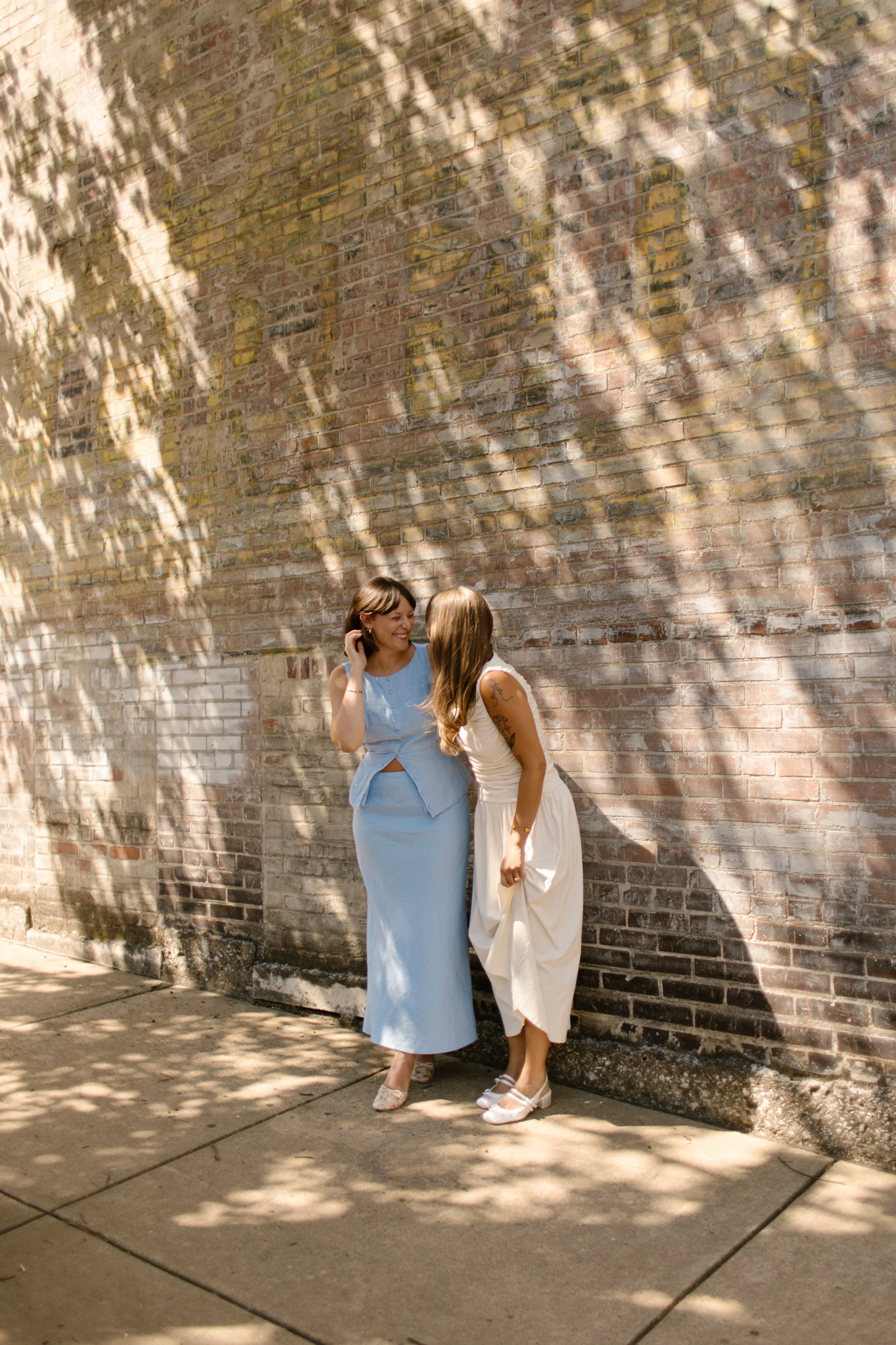 Two brides share a playful moment beside a sunlit brick wall, photographed by a documentary wedding photographer.