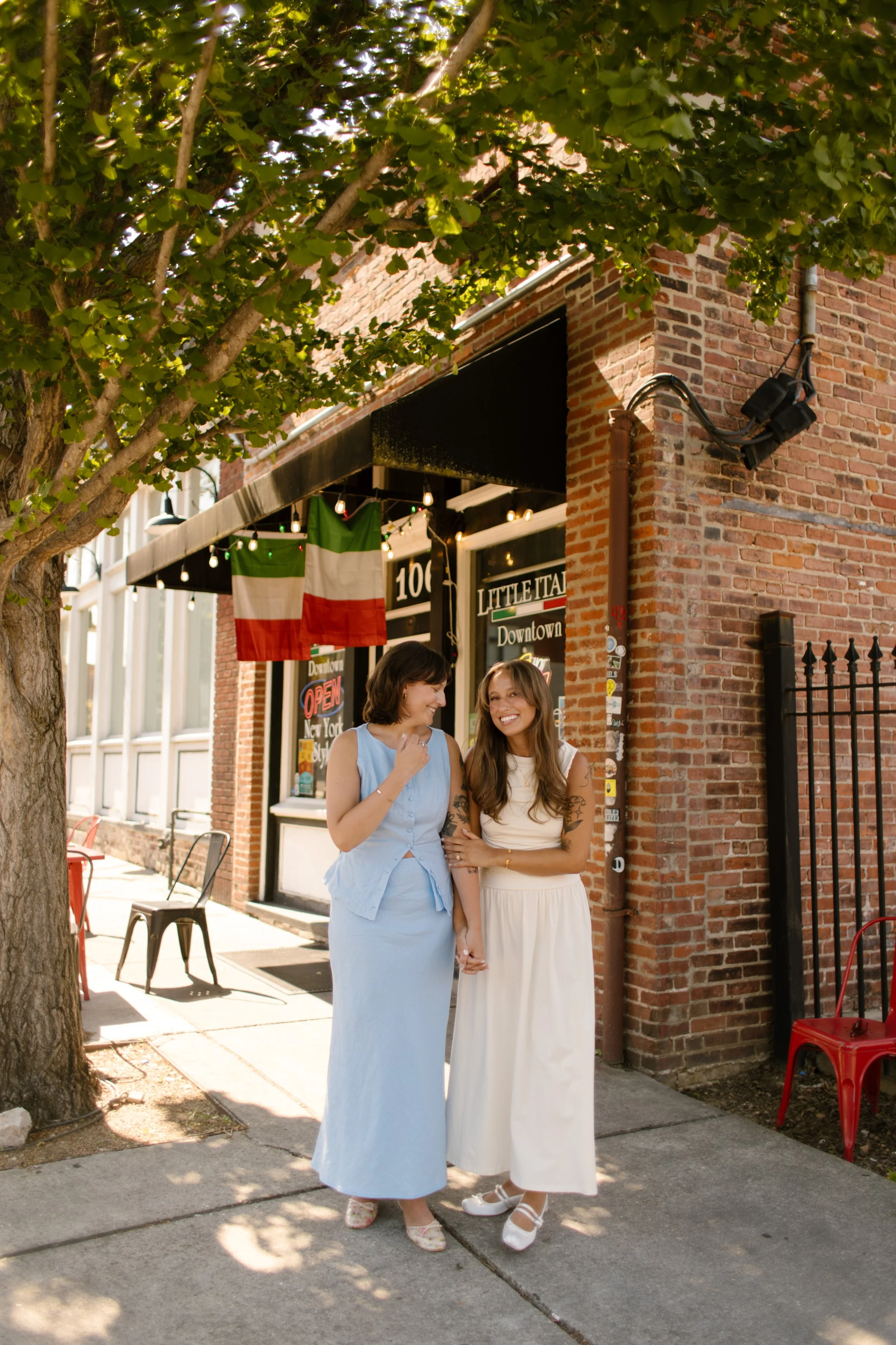 Two newlyweds smile together outside a neighborhood restaurant on a sunny downtown street.