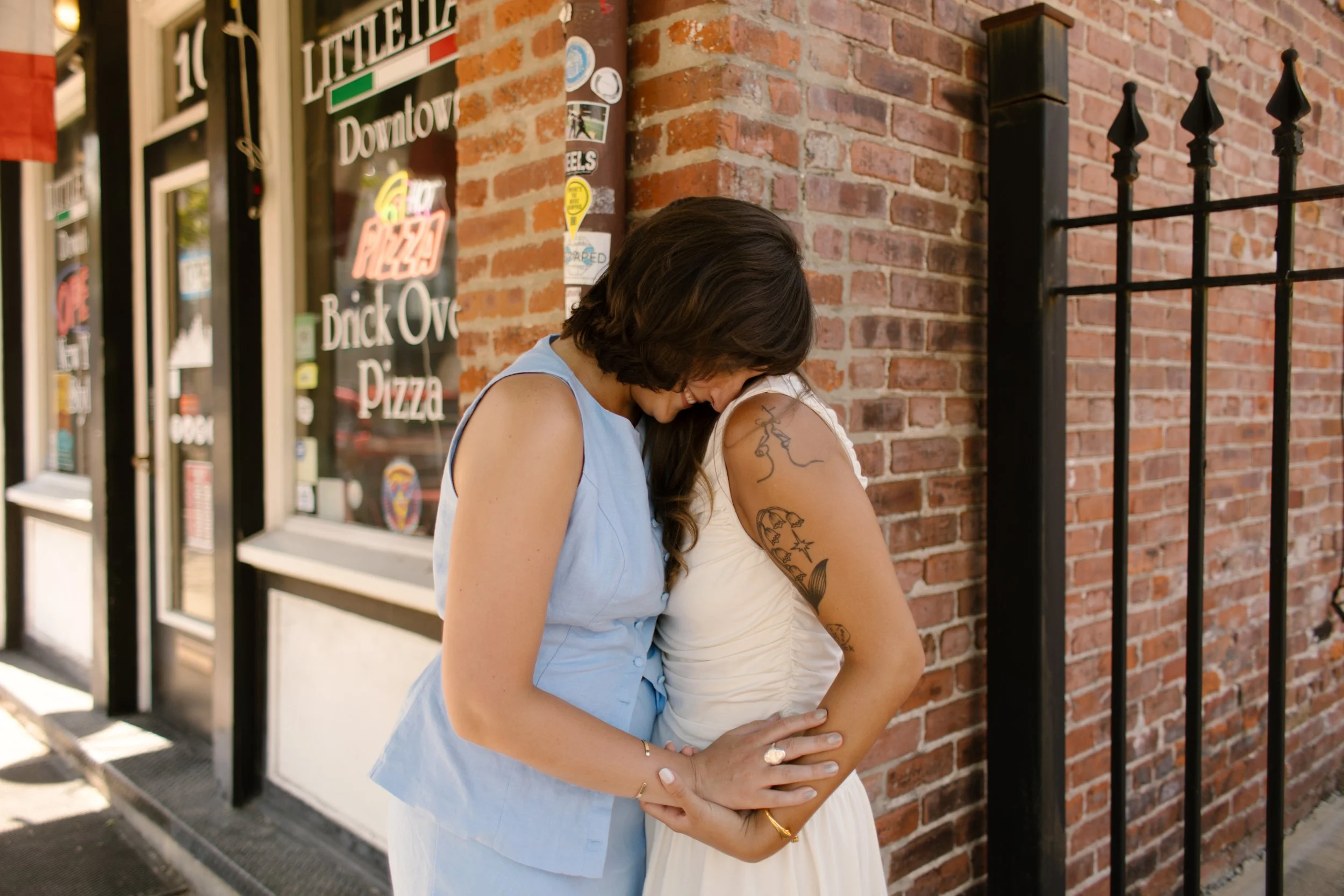 One bride rests her head against her partner outside a brick storefront in a candid wedding portrait by a documentary wedding photographer.