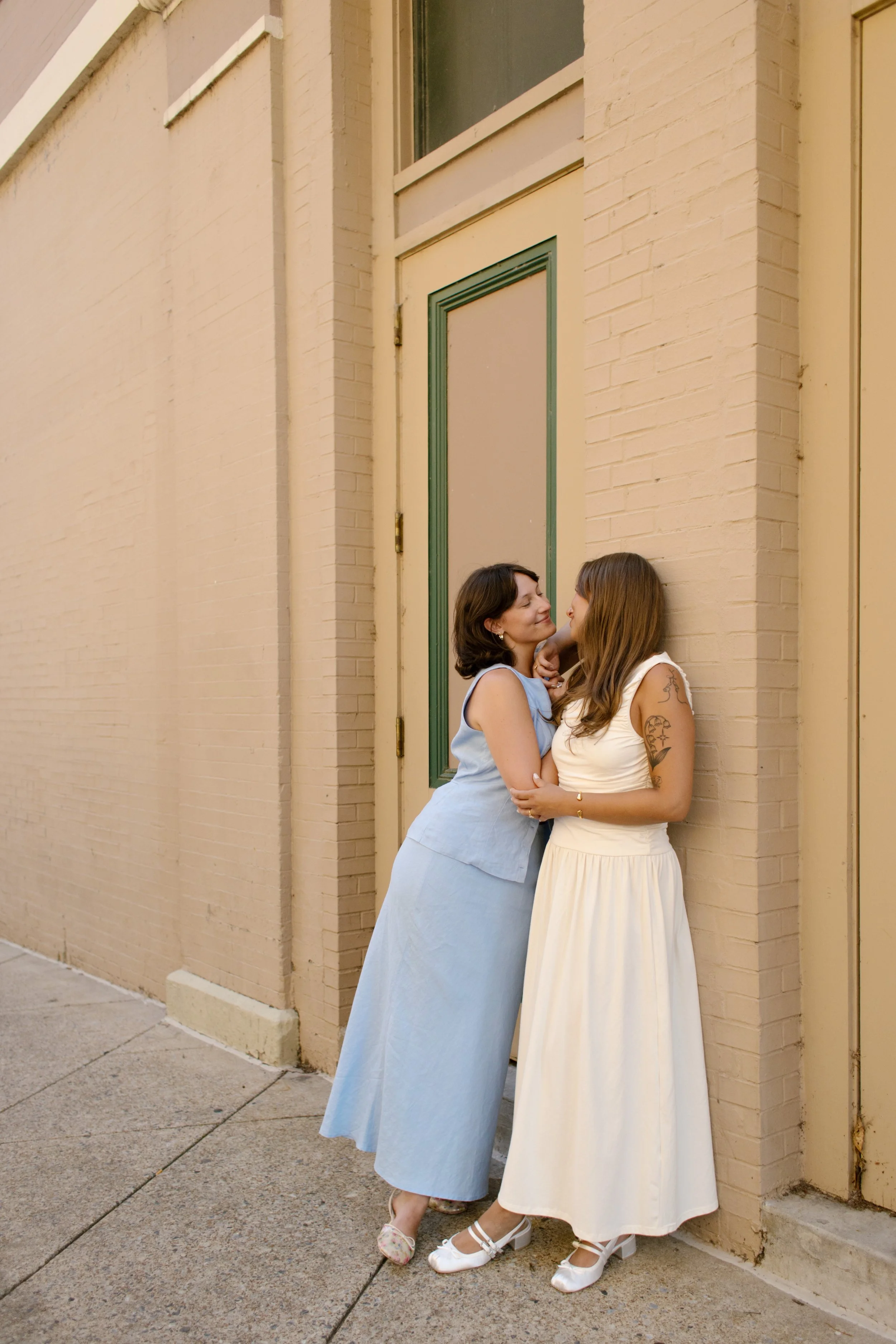 One bride kisses her partner's cheek while standing against a cream-colored building, photographed by a documentary wedding photographer.