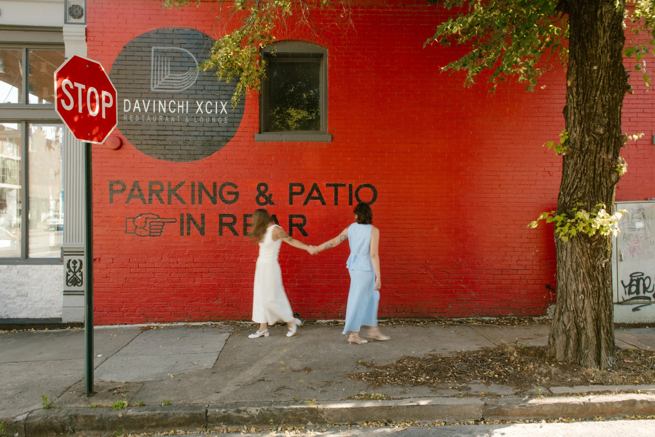 Wide shot of two brides holding hands in front of a bold red brick wall and city sidewalk.