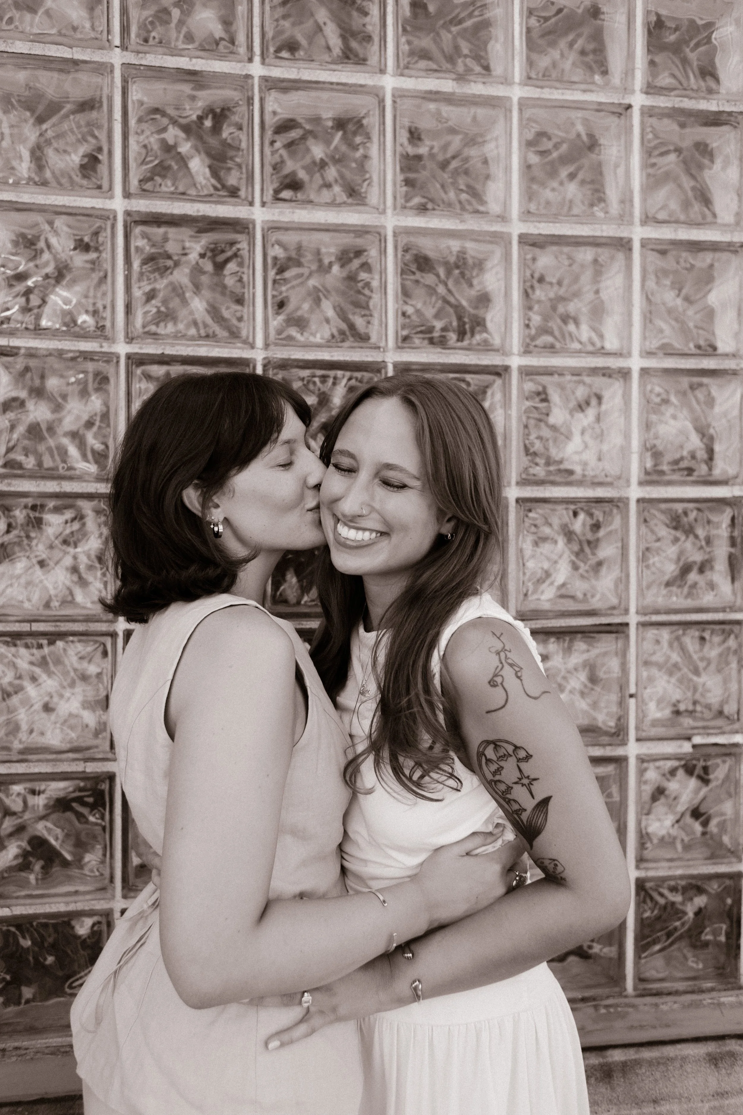 Black-and-white portrait of one bride kissing her partner's cheek in front of a glass block wall, captured by a documentary wedding photographer.