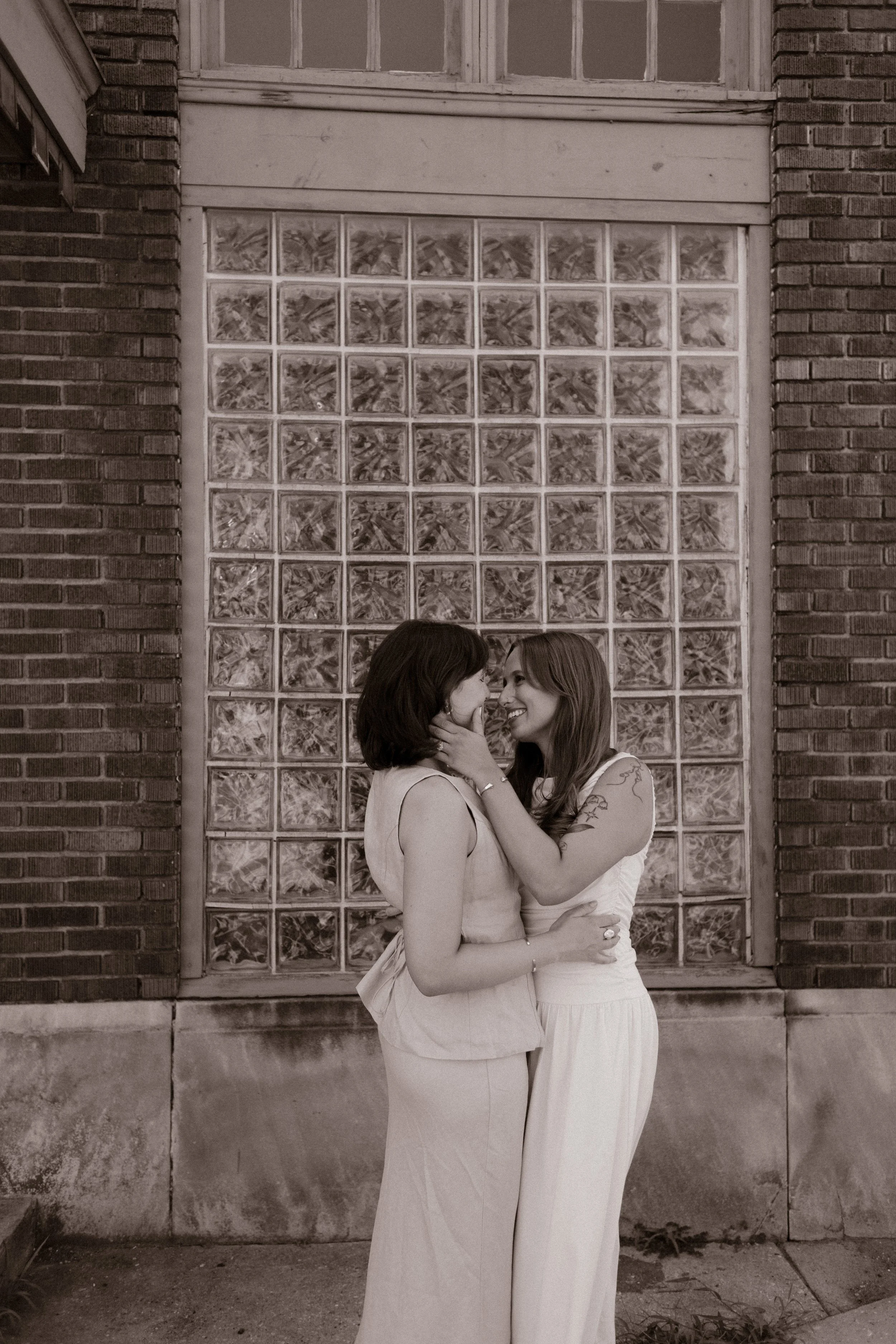 Black-and-white portrait of two newlyweds embracing in front of a glass block wall during their wedding day.