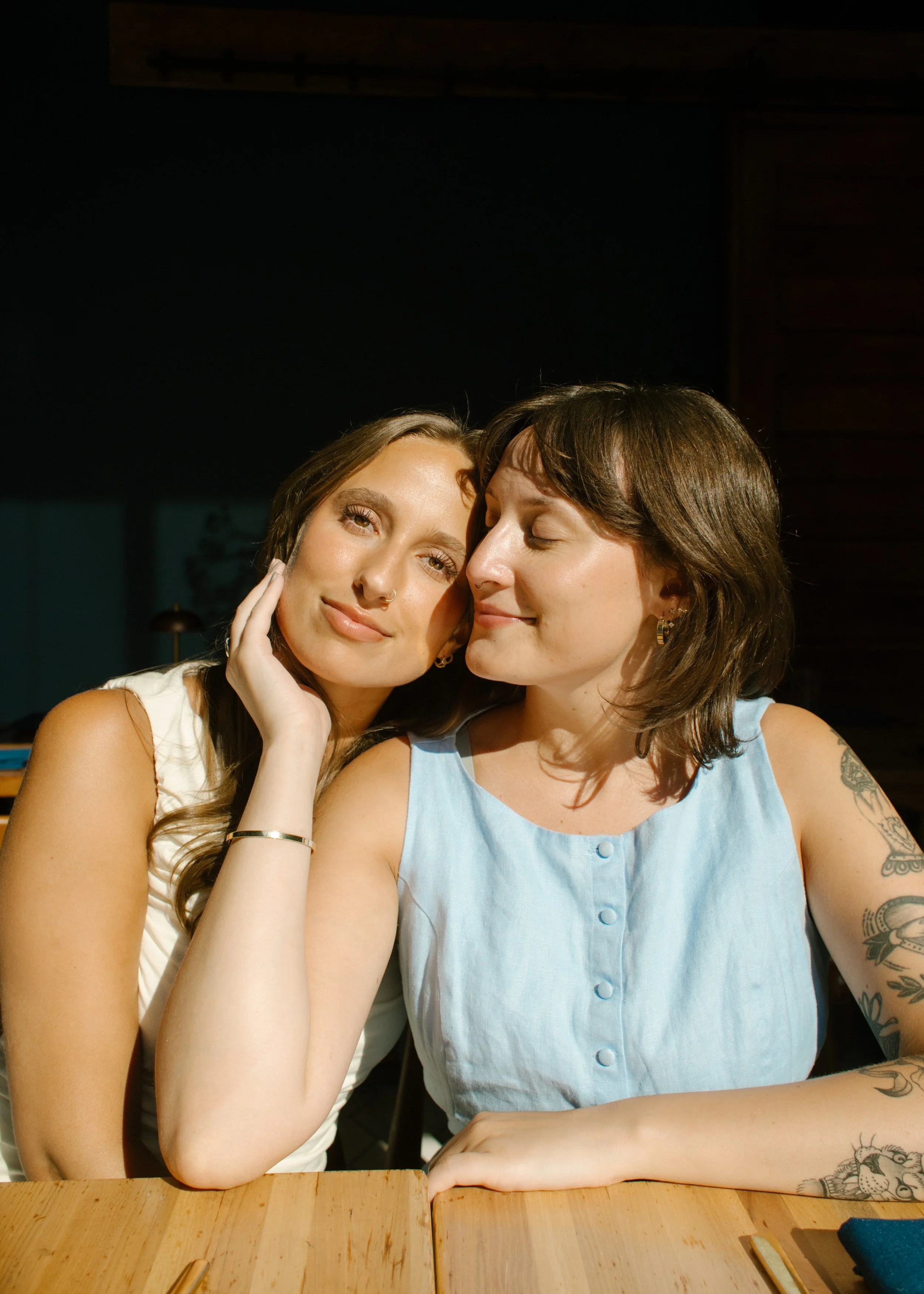 Sunlit portrait of two brides seated together at a wooden table during their wedding day, photographed by a documentary wedding photographer.