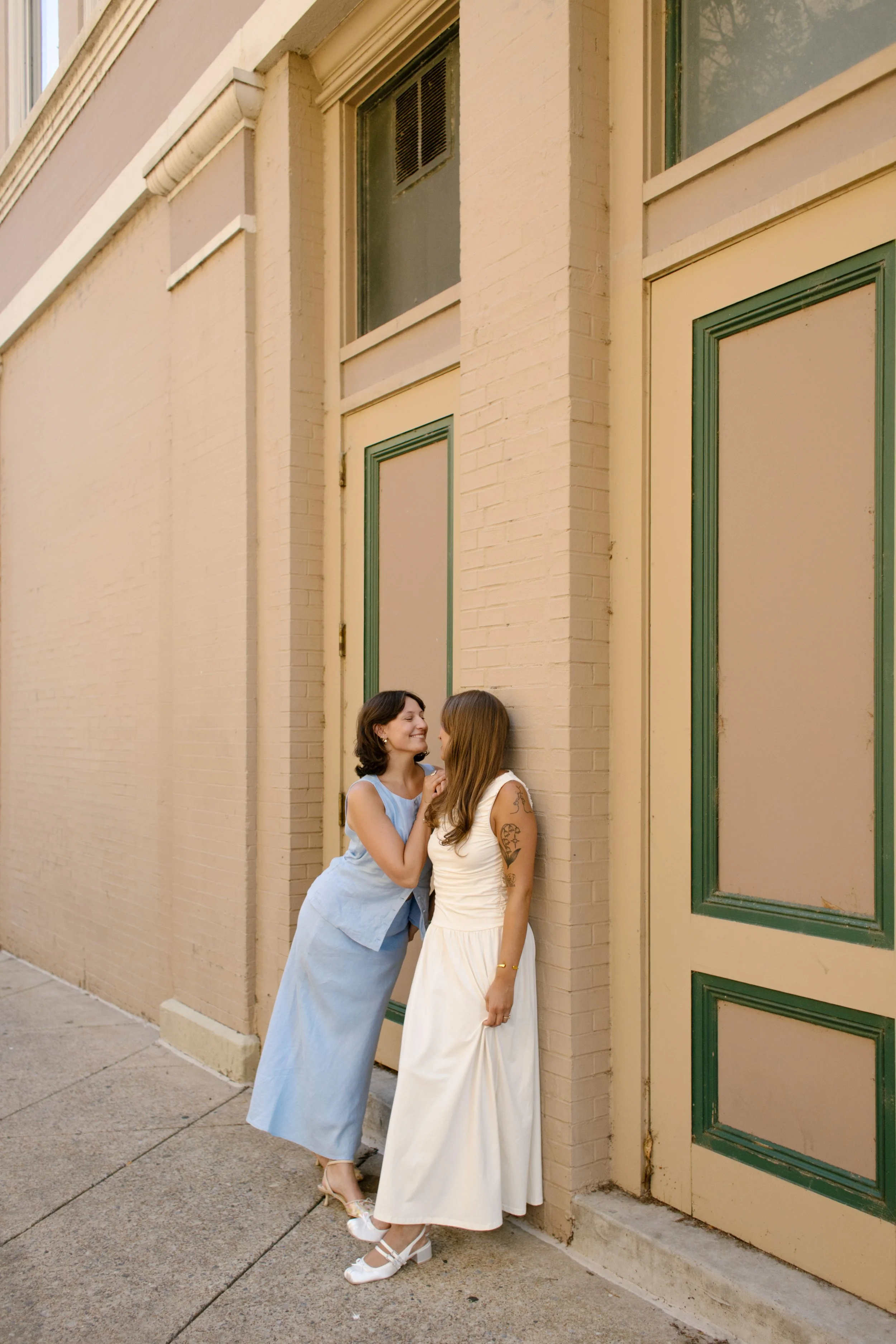 Two brides laugh together against a cream-colored building during an intimate city portrait session.