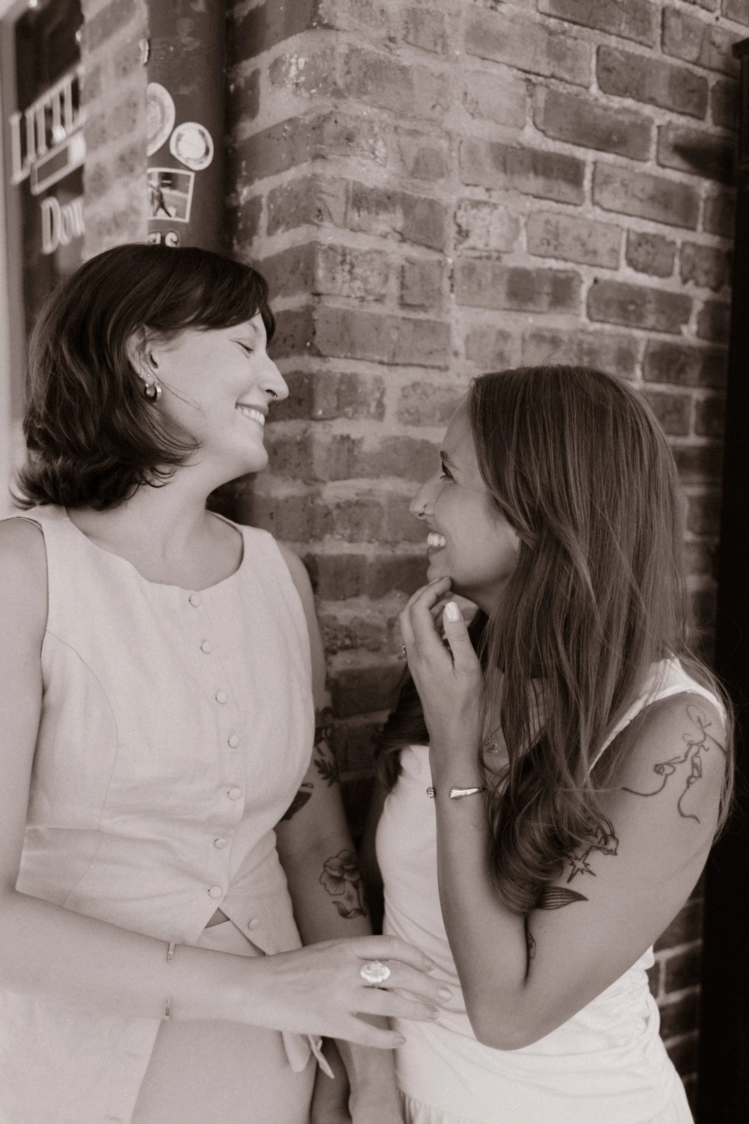 Black-and-white portrait of two brides smiling together outside a brick storefront, captured by a documentary wedding photographer.