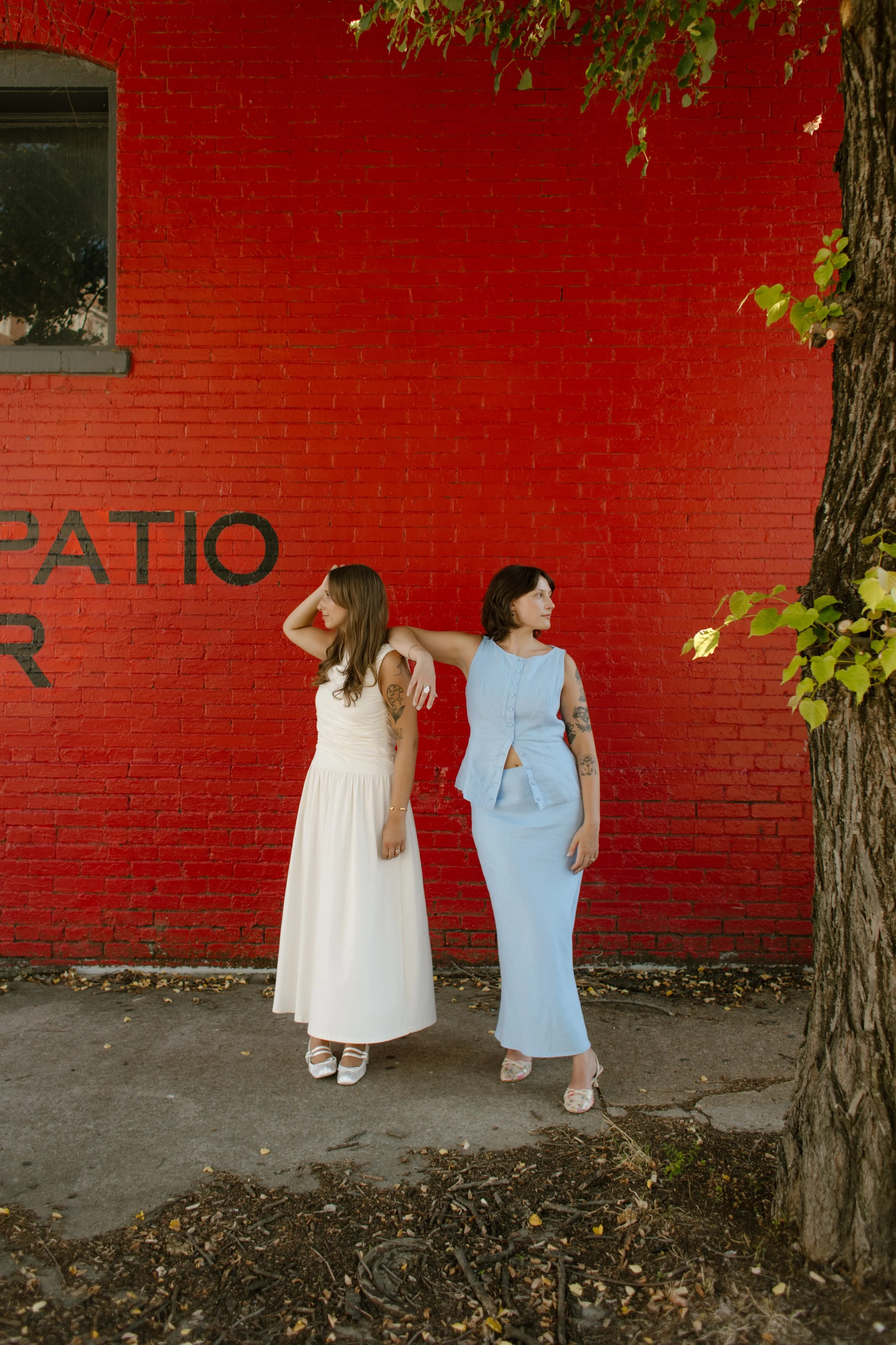 Two brides pose against a vibrant red wall beneath leafy trees during relaxed wedding portraits.