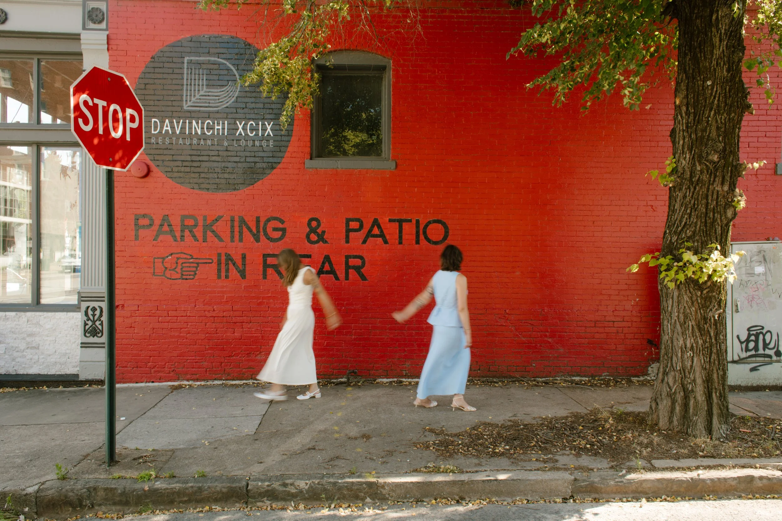 Wide shot of two brides walking past a bright red mural on a downtown sidewalk.