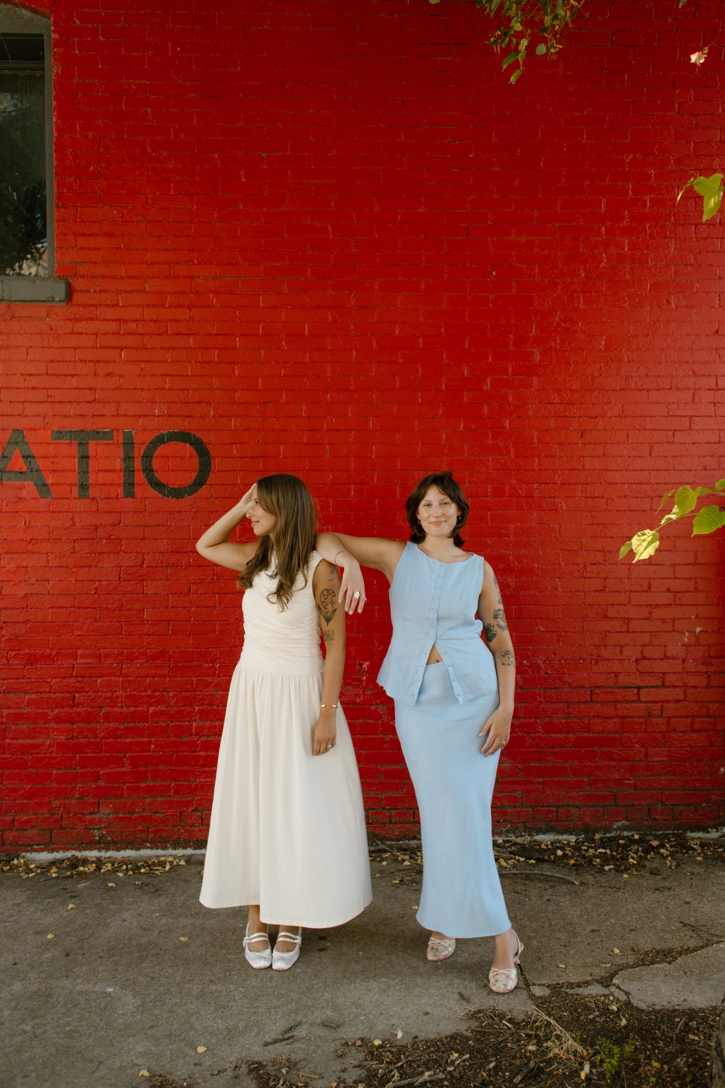 Two brides pose against a bold red brick wall in a colorful city portrait by a documentary wedding photographer.