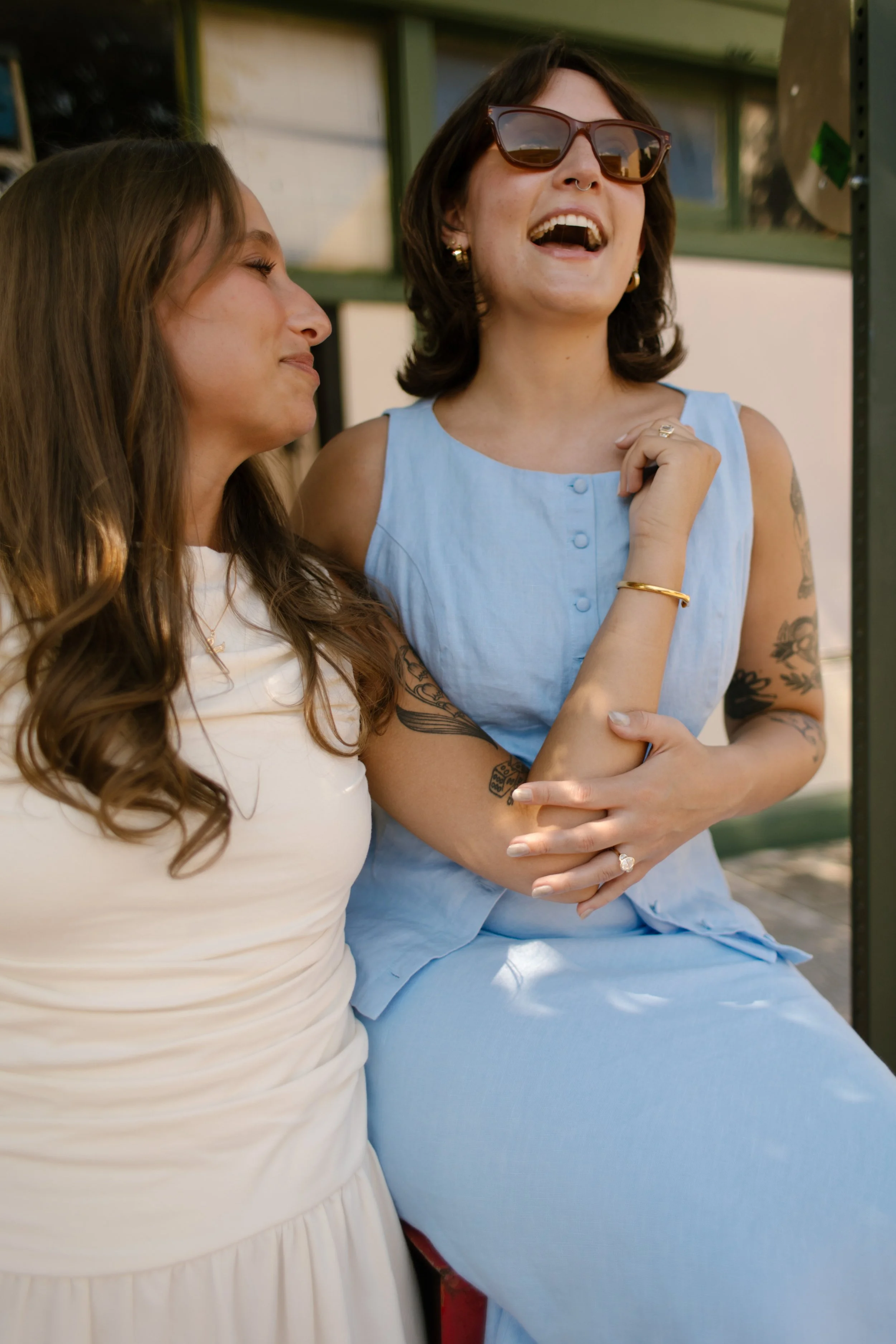 Two brides laugh together while sitting outside a storefront, captured by a documentary wedding photographer.