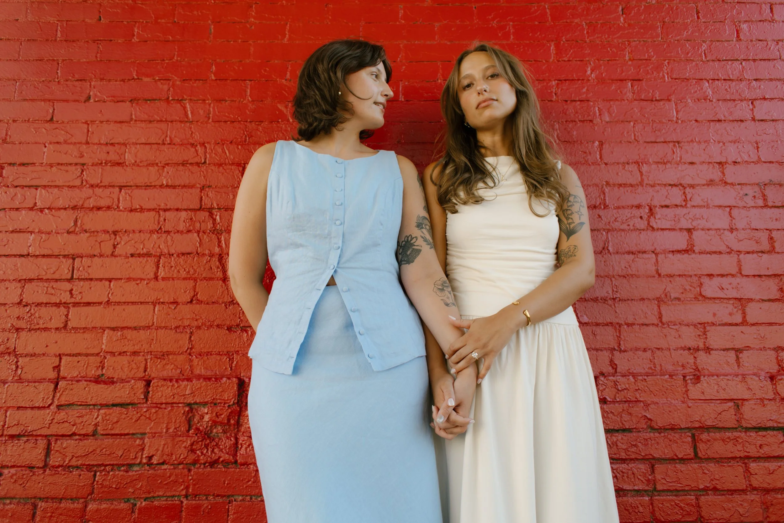 Two brides hold hands in front of a red brick wall for a modern city portrait.